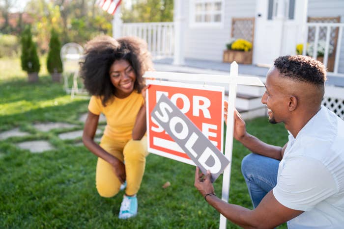 Young couple outside a house putting a &quot;Sold&quot; sign over the &quot;For Sale&quot; sign