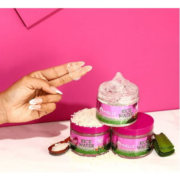 A model with the product on her fingertips with three product jars, a pile of rice and aloe vera with a magenta backdrop.