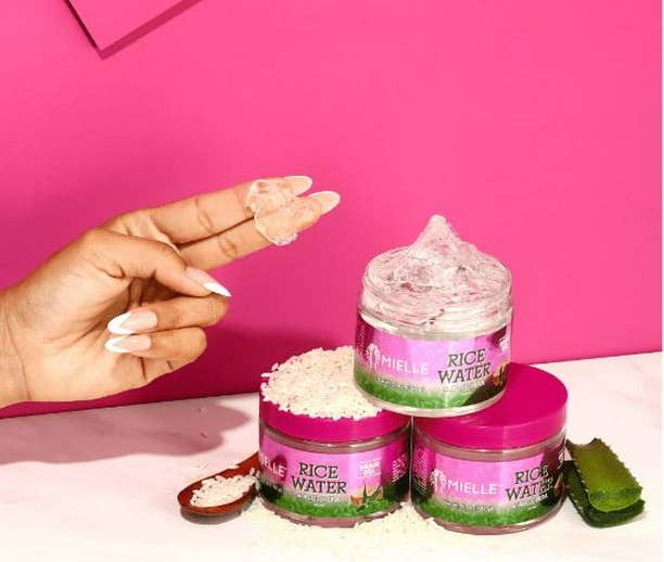 A model with the product on her fingertips with three product jars, a pile of rice and aloe vera with a magenta backdrop.