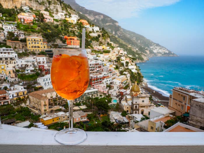 A bright orange Aperol Spritz cocktail resting on a ledge overlooking a quaint clifftop town surrounded by bright blue ocean