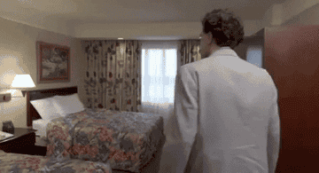 A GIF of Borat walking into a drab hotel room with floral bedsheets and curtains