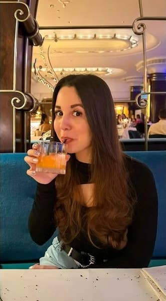 An image of Rhianna. She is wearing a long sleeved black top and drinking an orange juice drink from a straw. She has long dark brown hair which is wavy and falls to her waist.