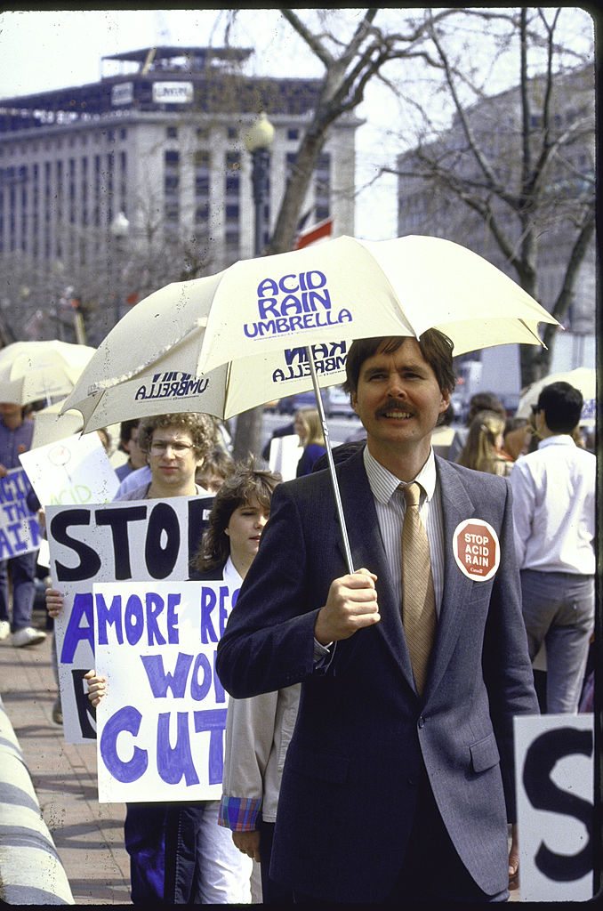A man holding an &quot;acid rain umbrella&quot; and wearing a &quot;stop acid rain&quot; sticker in a crowd of protesters