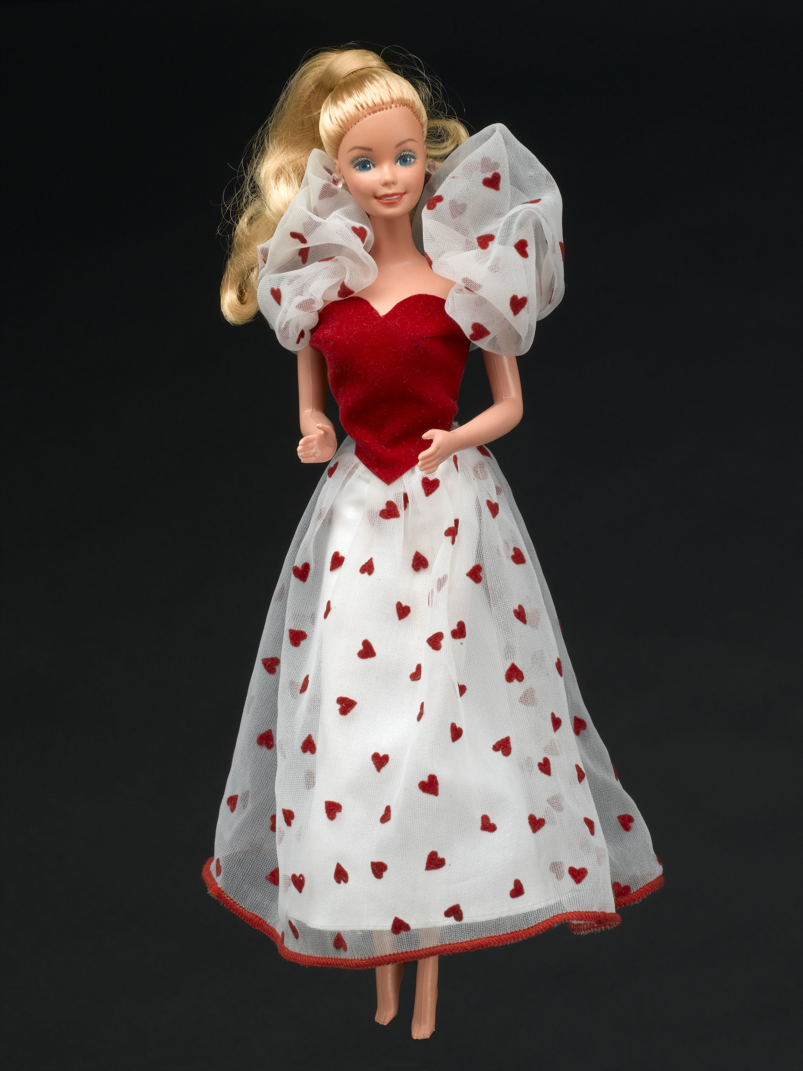 Barbie doll, by Mattel, 1980s. Plastic doll, white skinned, with shoulder-length blonde hair, in long skirt of hearts pattern, and red sleeveless top and white tulle stole with hearts