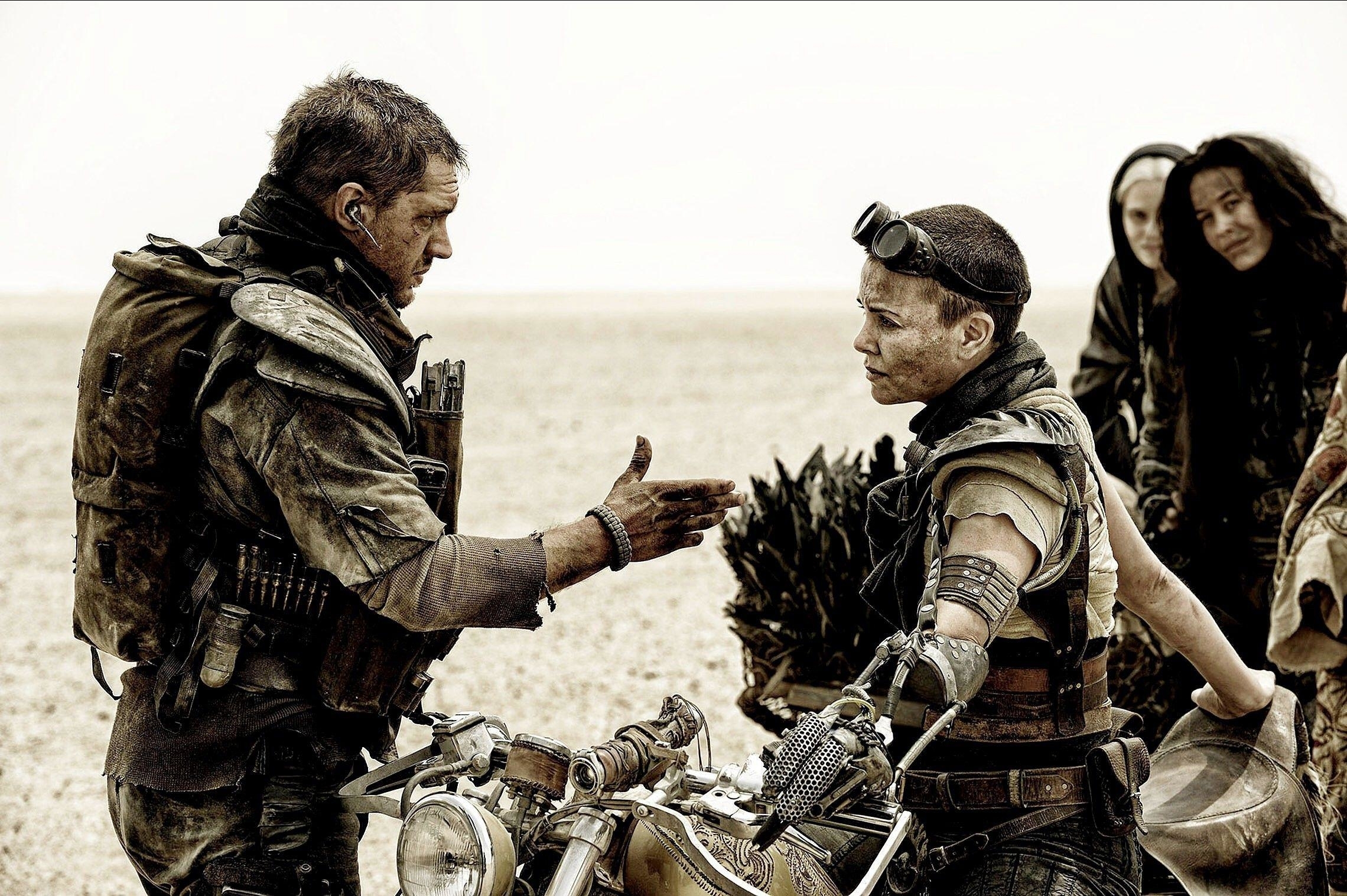 Tom Hardy extends a handshake to Charlize Theron in a dusty desert