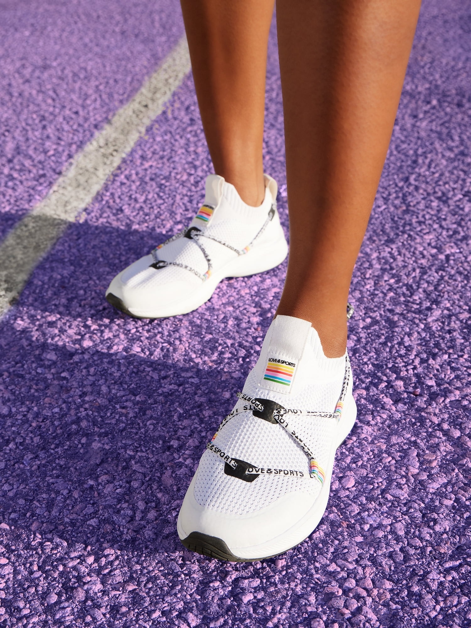 a model wearing the white, black and rainbow sneakers on a purple tennis court