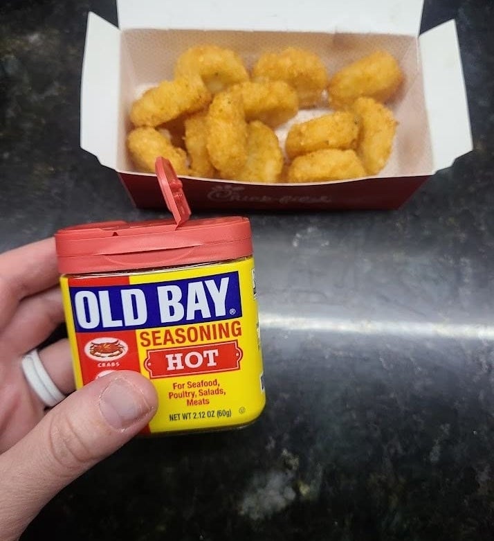 Reviewer&#x27;s photo of them holding a small container of Old Bay seasoning in front of a box of tater tots