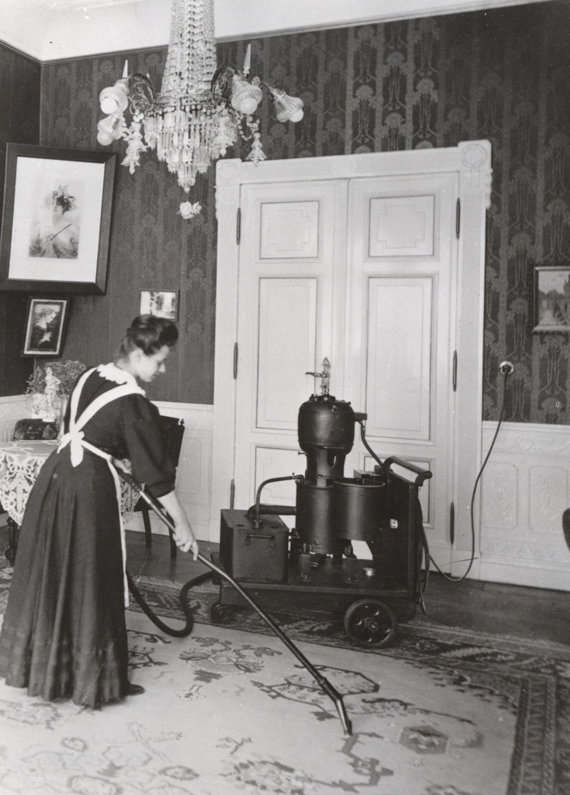 A woman in a long dress and apron vacuuming with a long attachment connected to what looks like a small steam engine