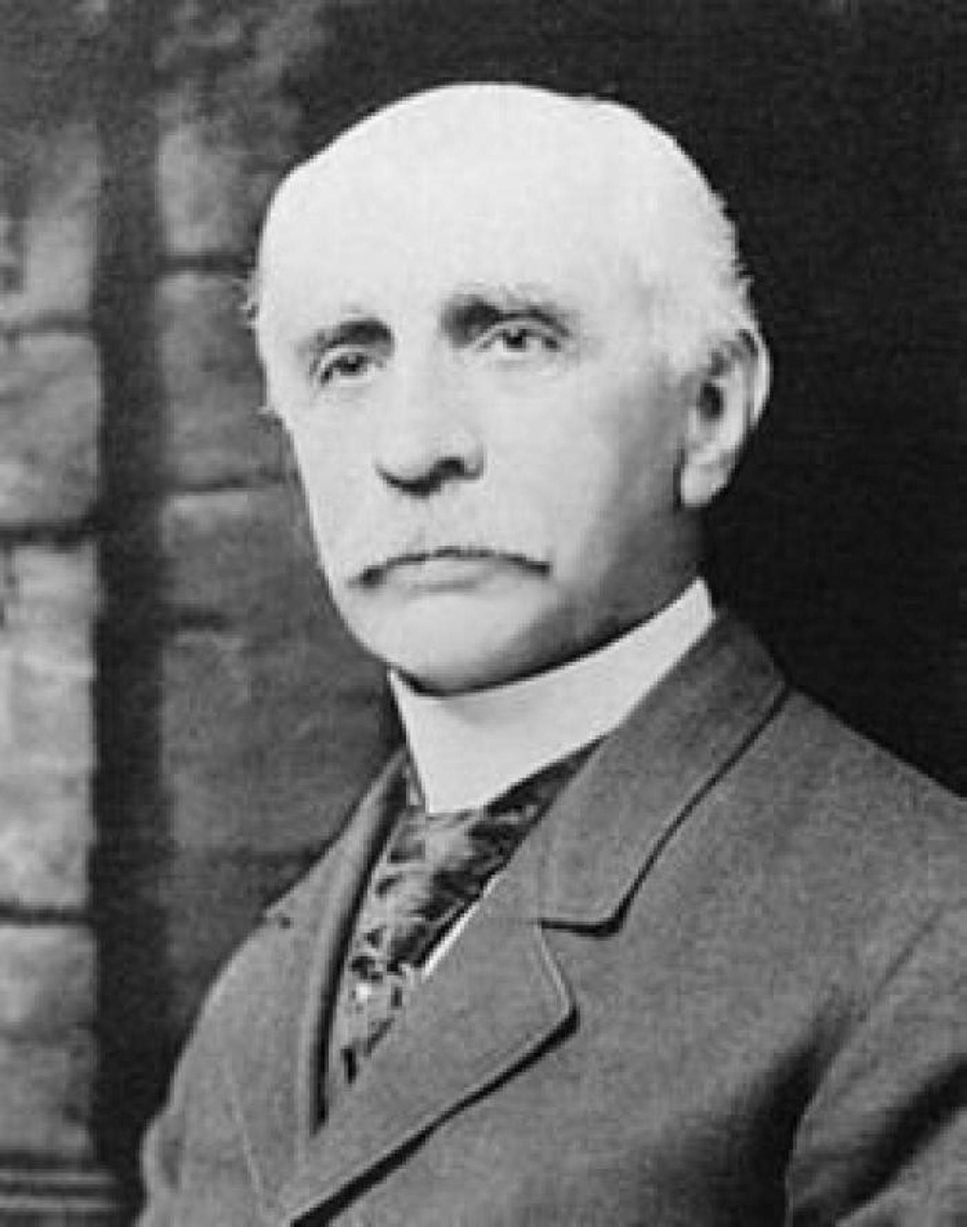 An unsmiling, balding man with white hair and a white mustache, wearing a suit and tie