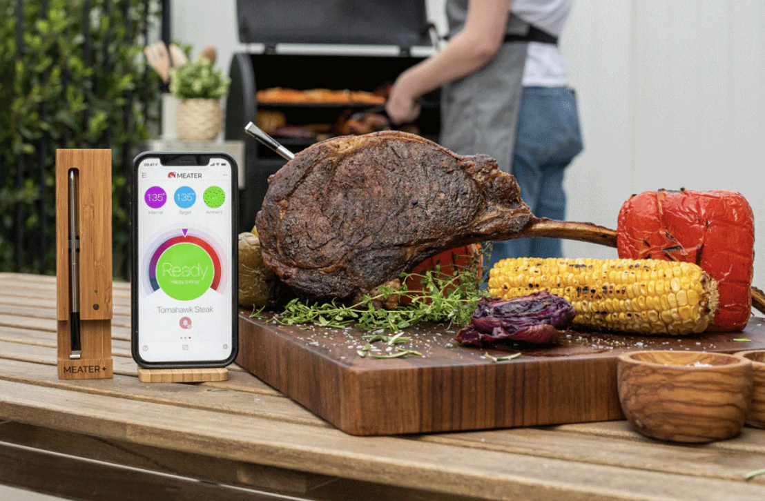 a meat thermometer inside a piece of meat next to an iphone, corn, and a pepper