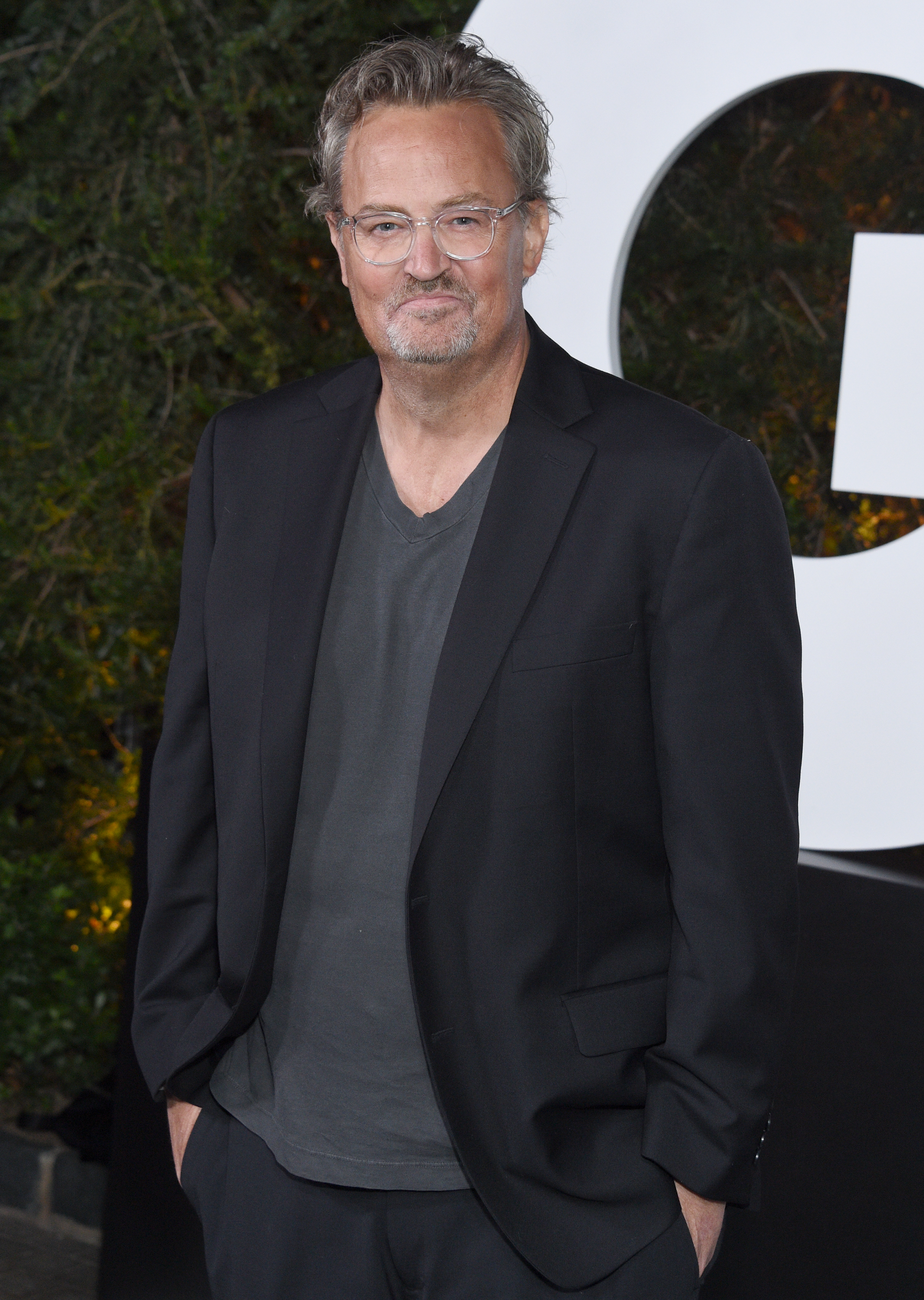 wearing a blazer over a t-shirt with glasses and grey hair combed back