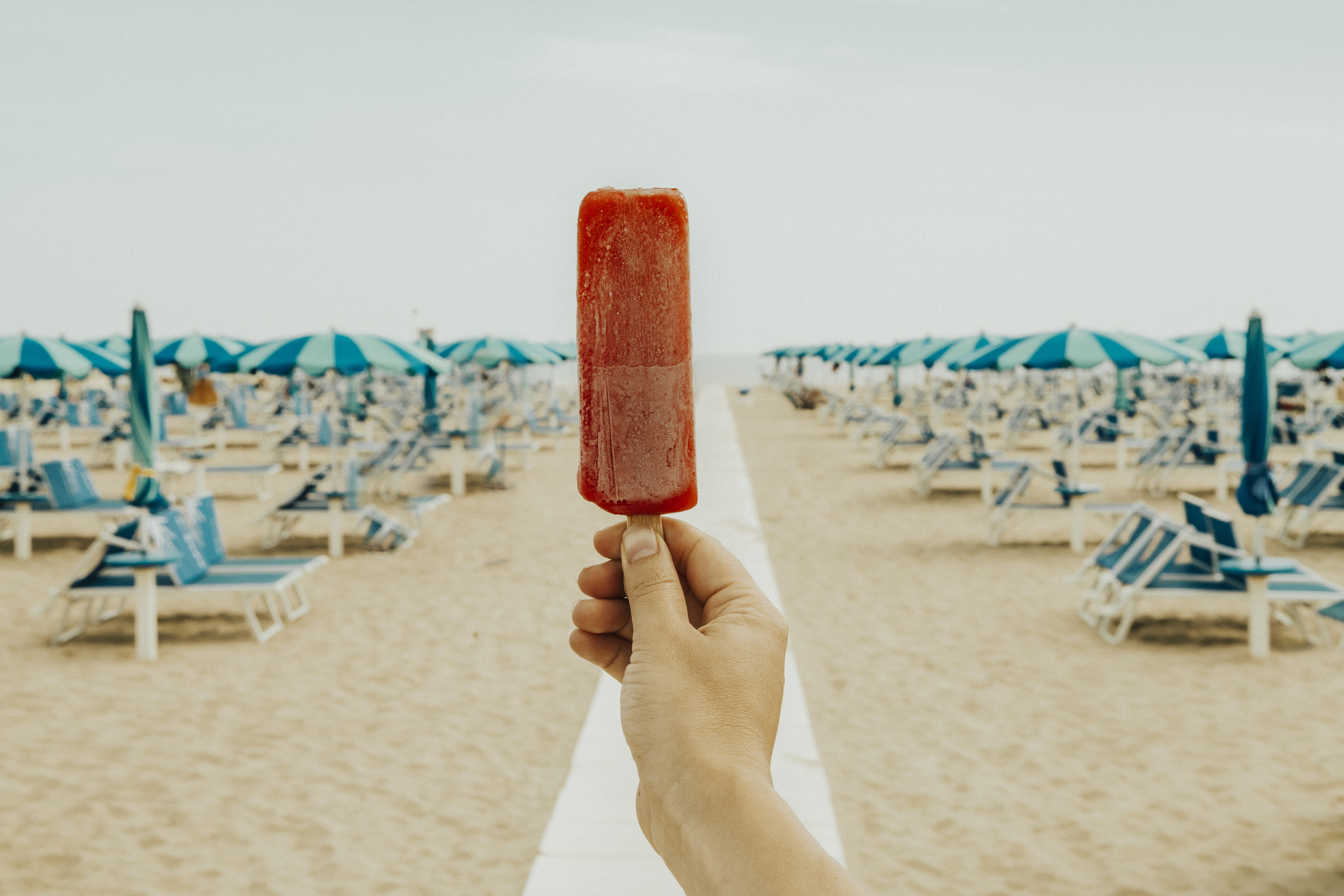Holding up a popsicle against a beach backdrop filled with lounge chairs and umbrellas