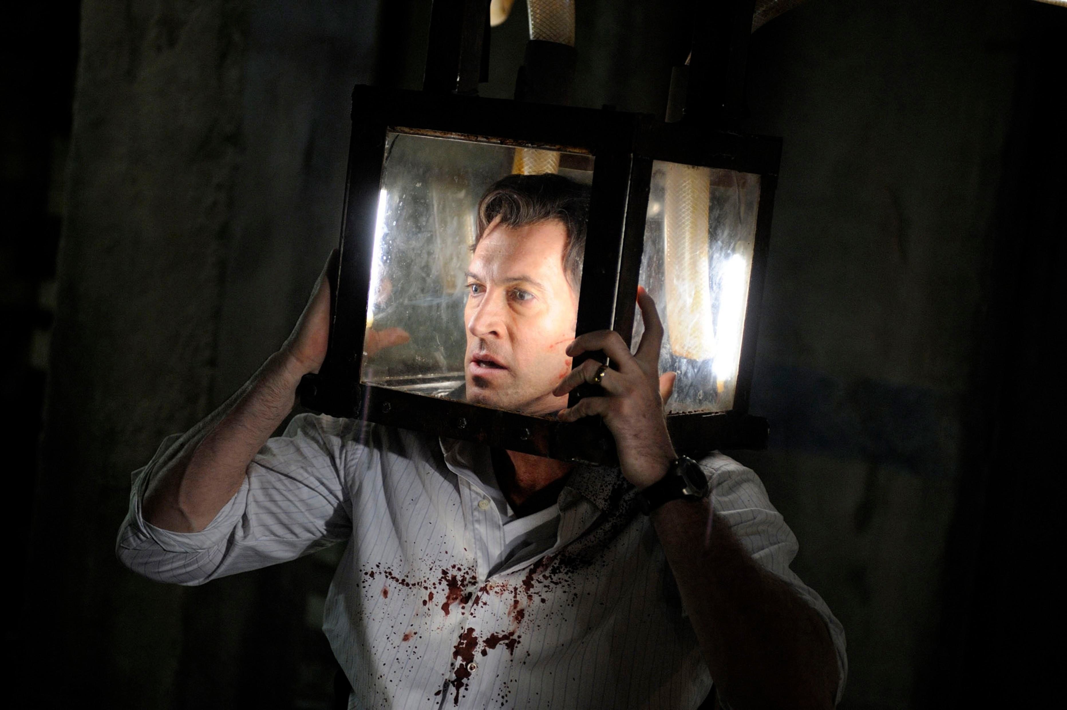 Scott Patterson, wearing a blood-stained shirt, stands with a glass box on his head.