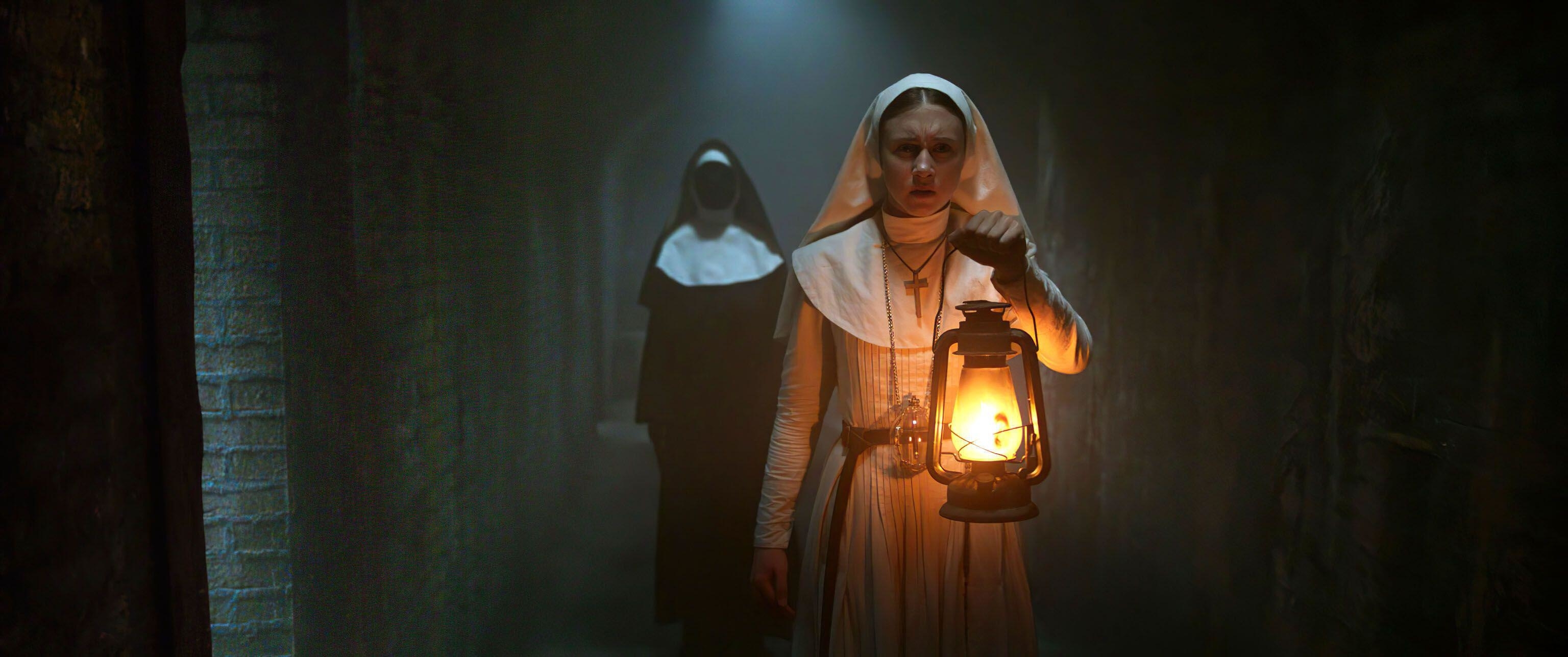 Taissa Farmiga walks with a lantern down a dark tunnel with a creepy nun following behind her