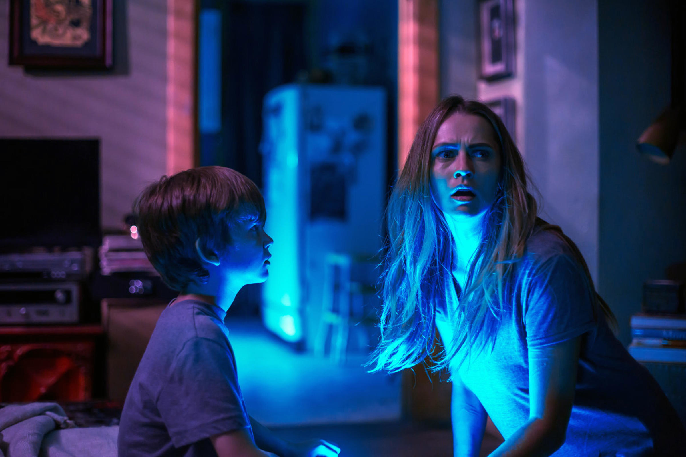 A young woman and a boy sit with great fear while basking in a bright blue light
