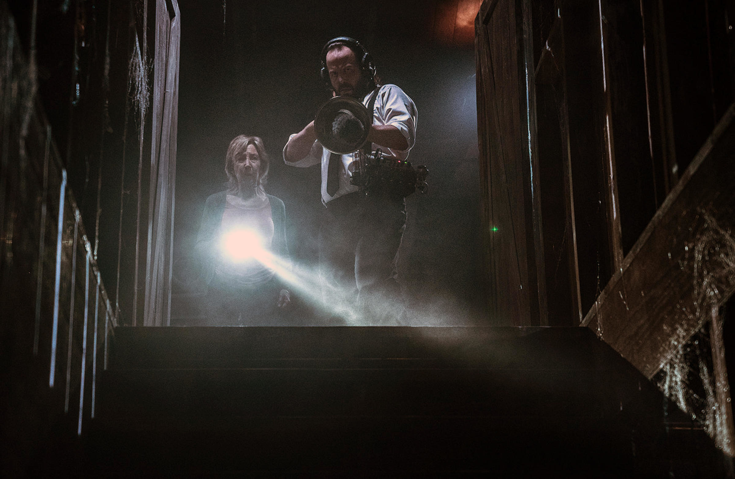 A man and woman hold up a paranormal reading device and a flashlight toward a dusty basement