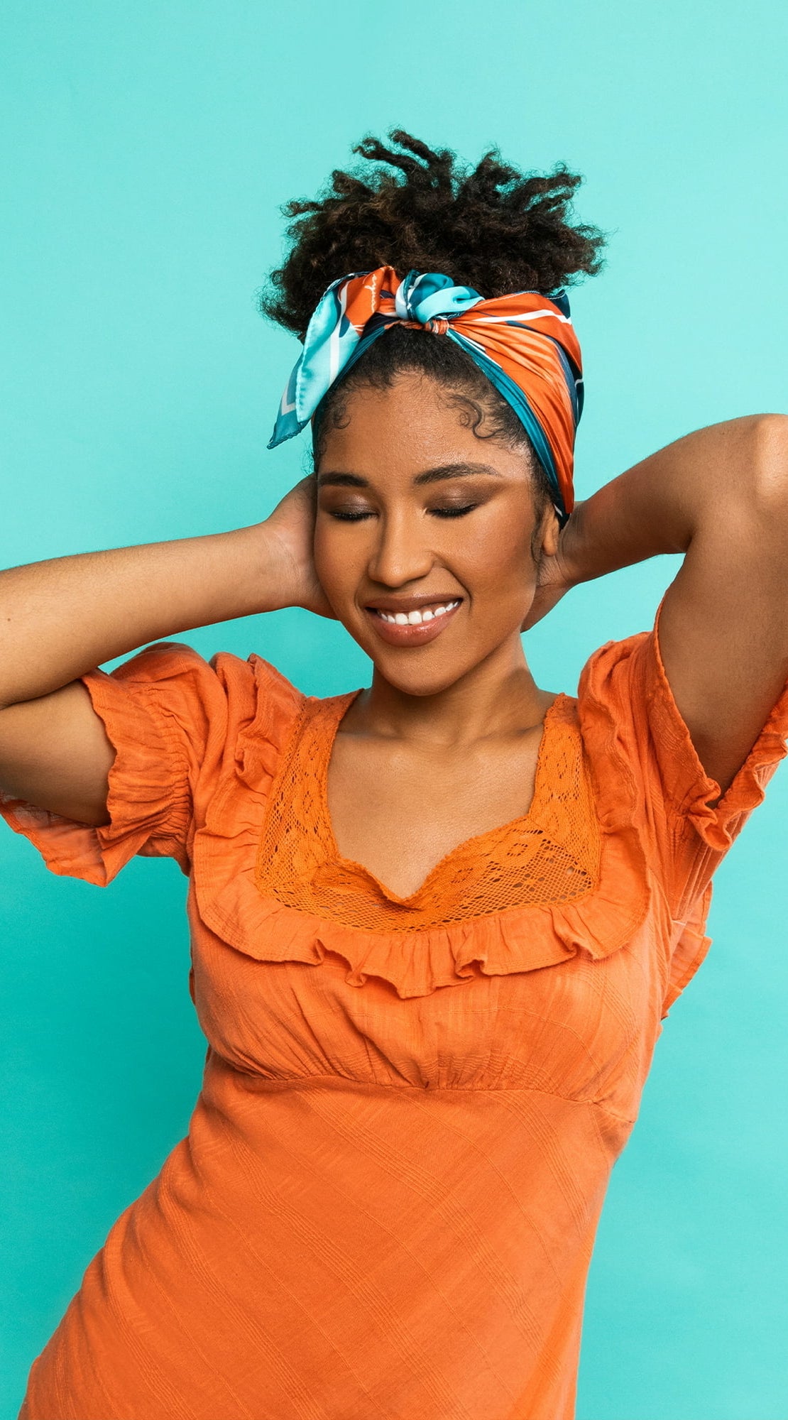 A model wearing the scarf wearing a bright orange dress with a teal backdrop