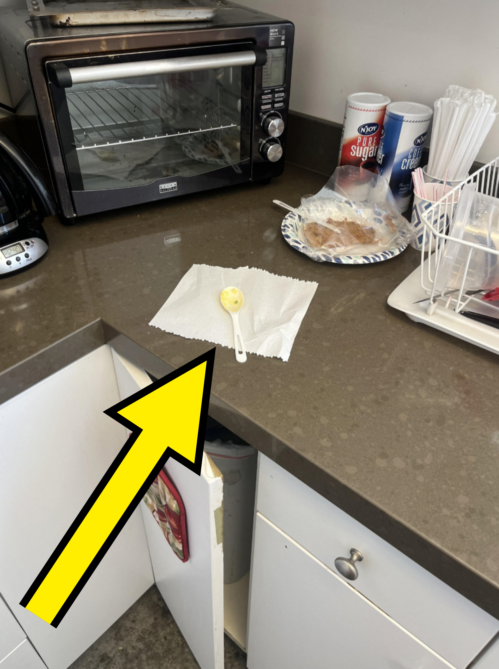 A used plastic spoon and paper towel have been left on the kitchen counter, directly above the open trash can