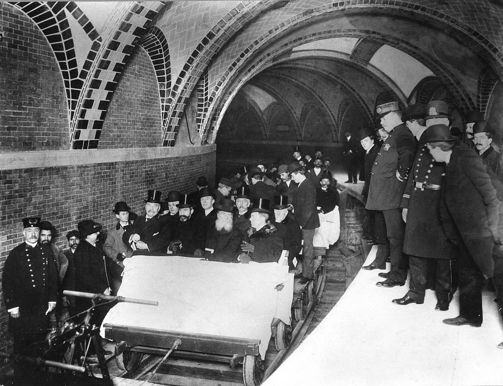 Men in top hats sitting in an open carriage and standing on the tracks and the platform