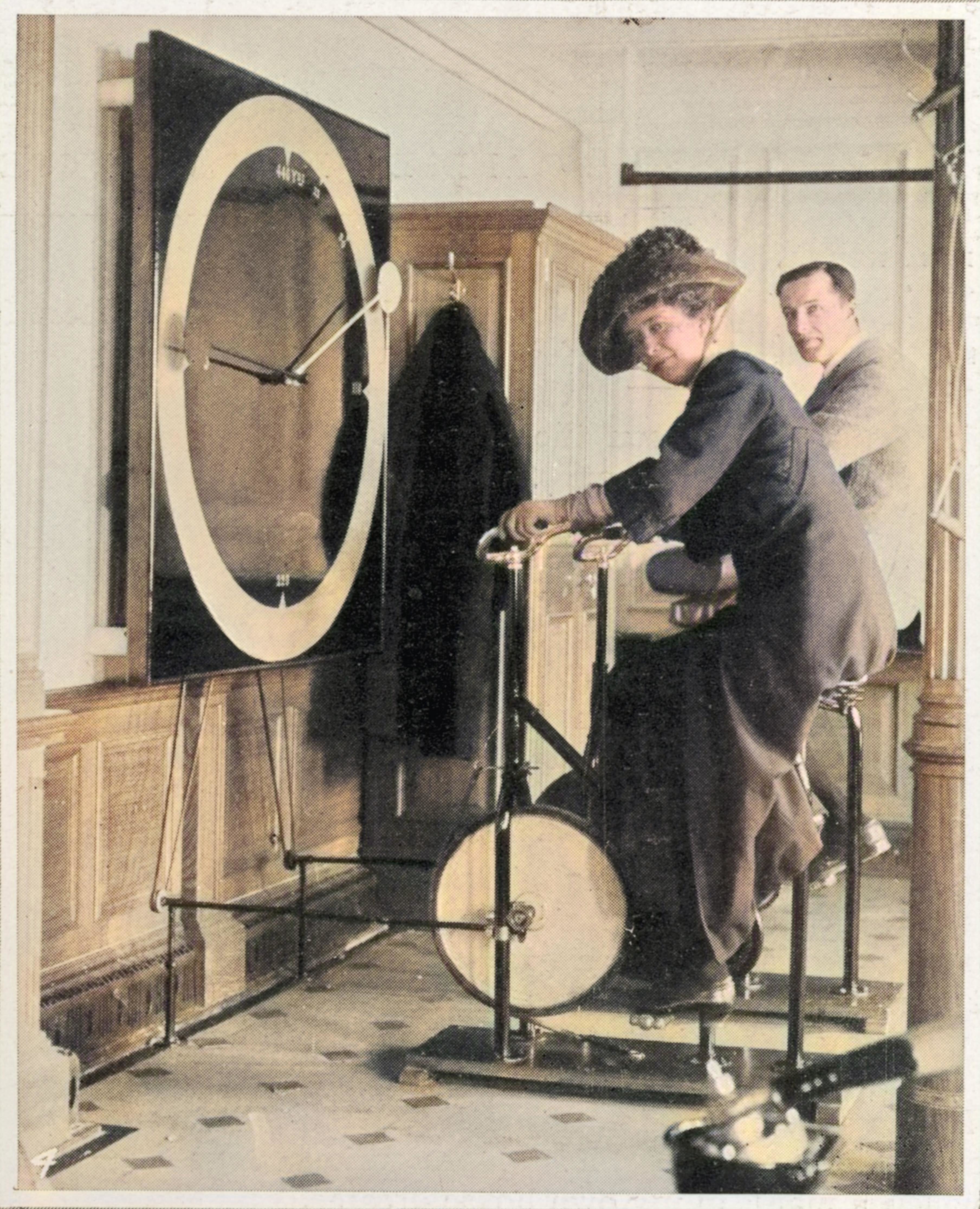 A woman wearing a long dress, coat, and ornate hat sits on an exercise bike next to a man also on a bike and looks at the camera
