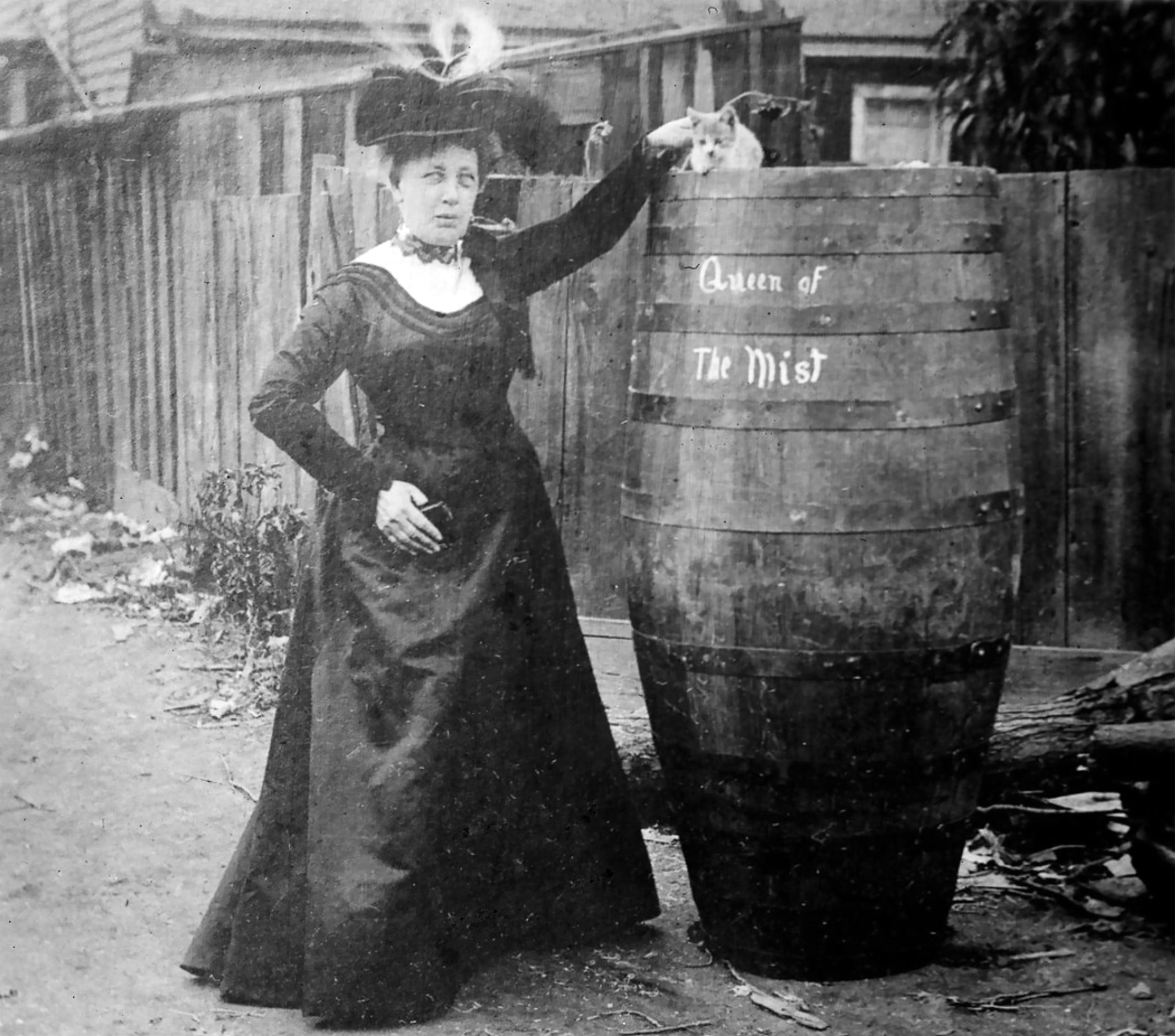 A woman in a high-necked, long dress with long sleeves and ornate hat standing next to a &quot;Queen of the Mist&quot; barrel