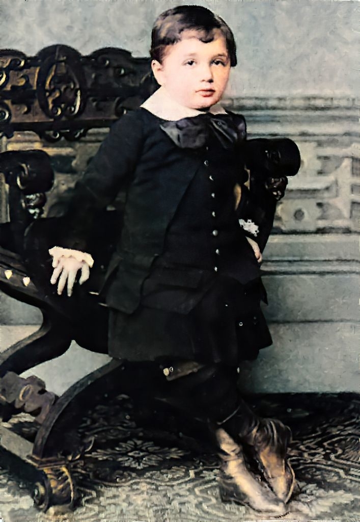 A serious-looking child with ankle boots, a dress suit, and tie leans on a chair