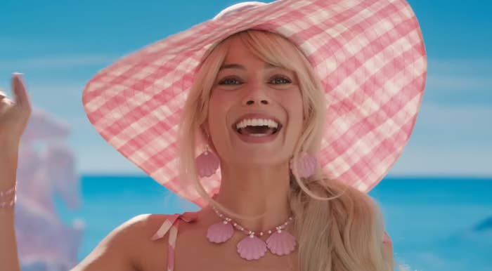 Margot Robbie as Barbie smiling and waving. Barbie is wearing a wide-brimmed plink plaid sun hat