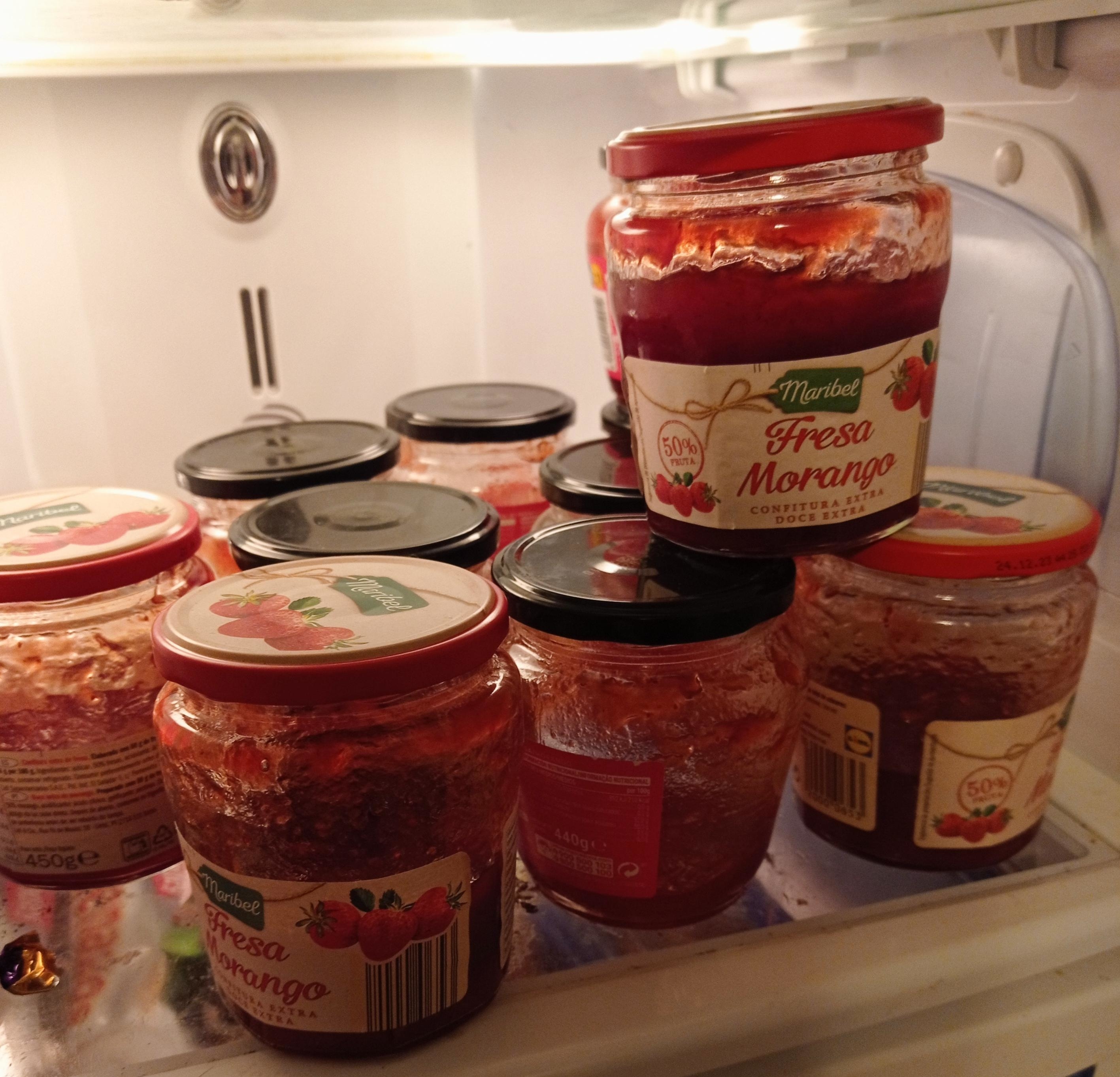 A shelf in the fridge with open jars of strawberry jelly
