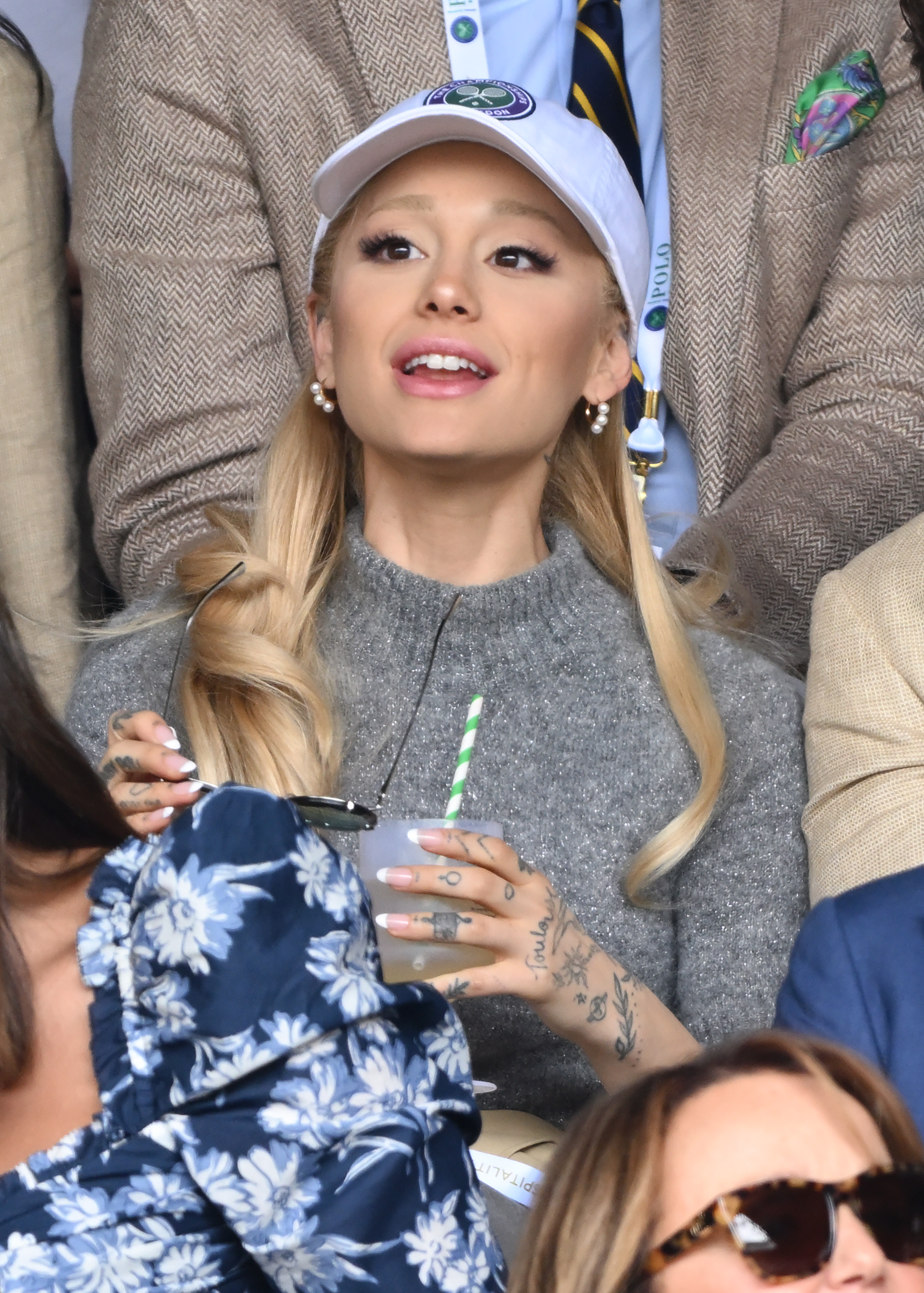 Close-up of Ariana sitting in an audience and wearing a cap