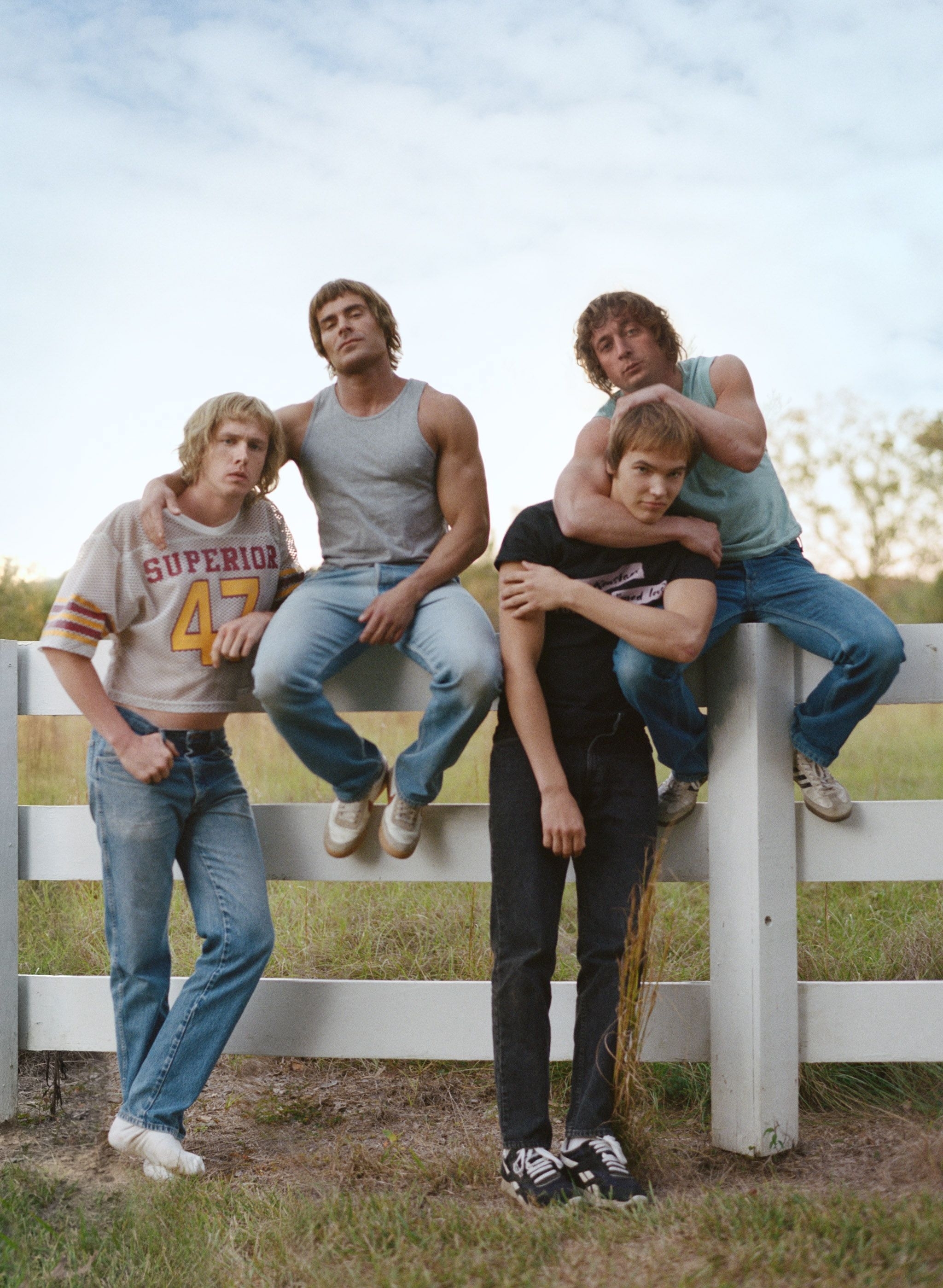 the four actors at a wooden fence wearing clothes from the 80s and sporting their shaggy bowl hair cuts