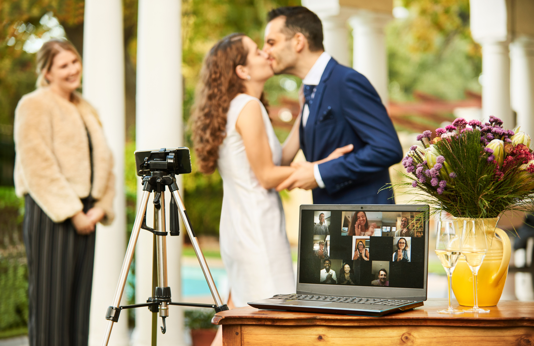 A couple kissing outdoors with a camera attached to a laptop showing Zoom attendees