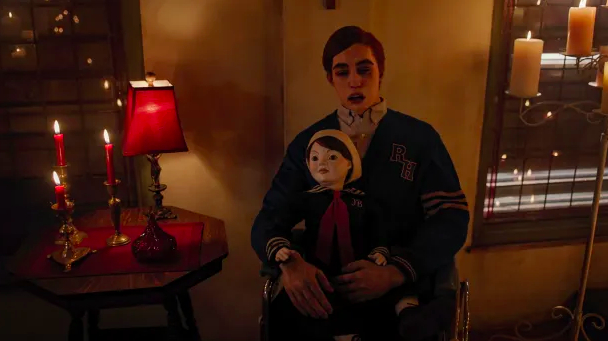 Jason&#x27;s dead body sits in a wheelchair. He has red hair, his eyes are closed and his mouth is open. He is wearing a shirt and cardigan. He is holding a creepy doll. The room is lit with candles.