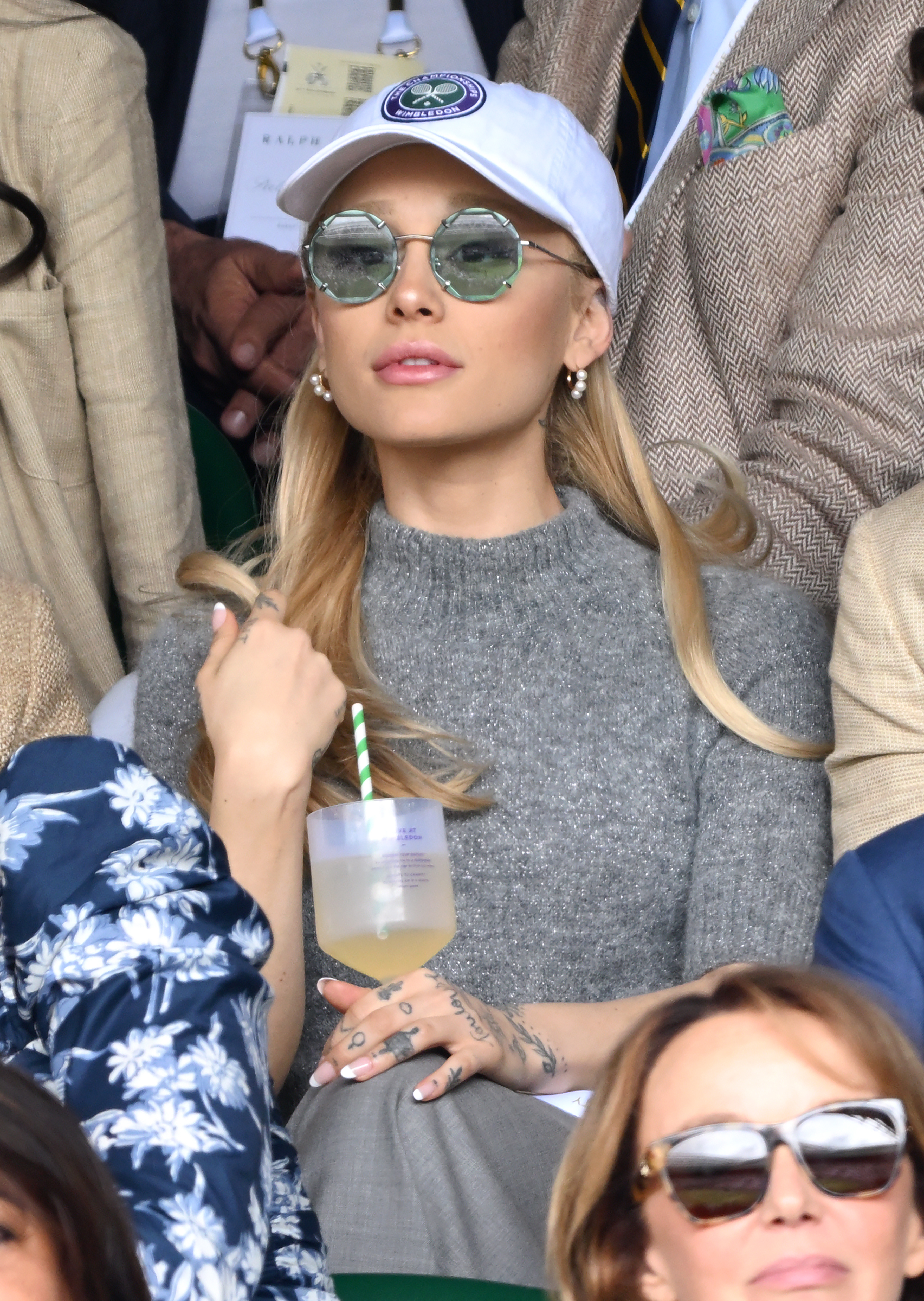 Ariana sits in the stands of a sporting event wearing a baseball cap and holding a cup