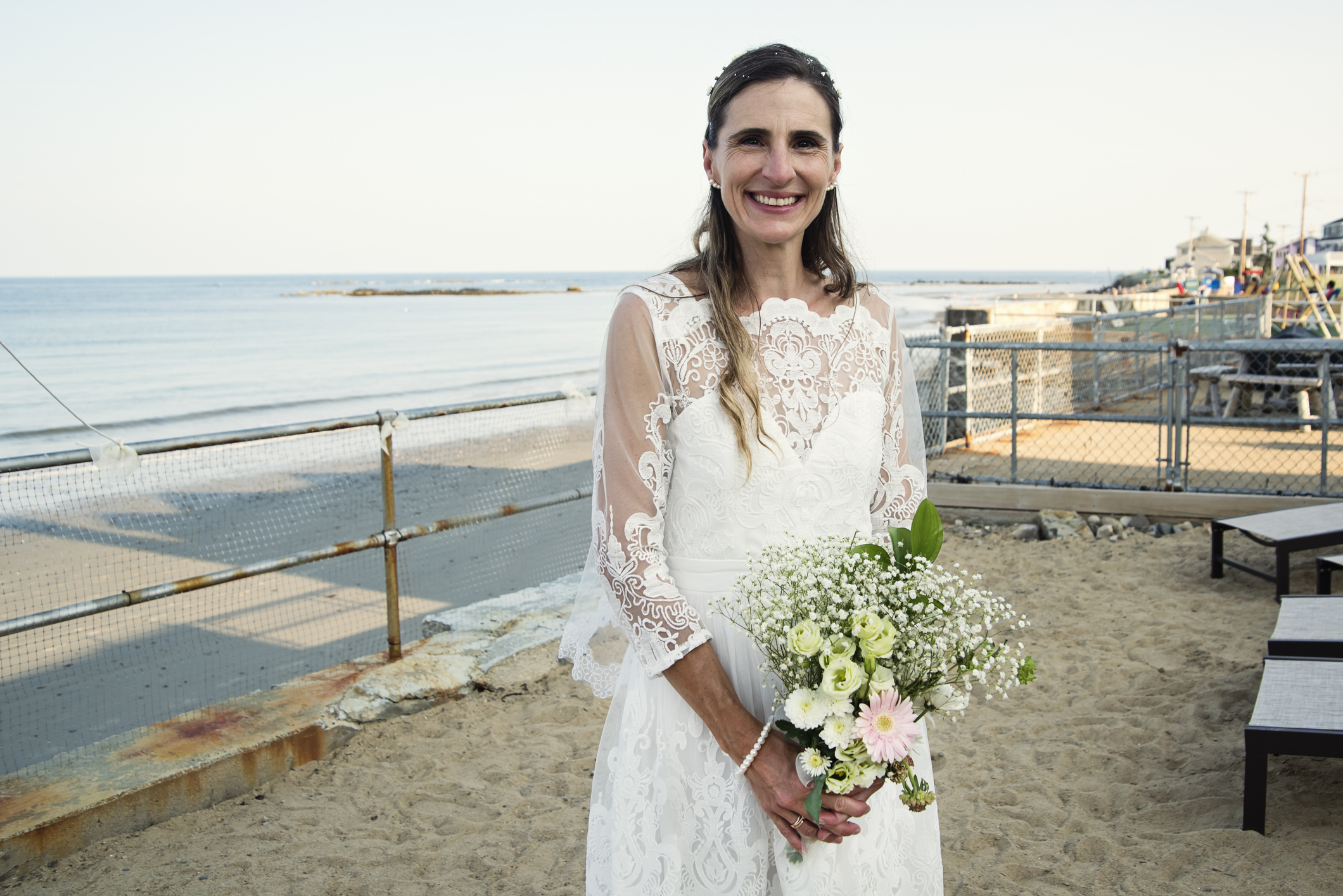 A woman in a bridal gown holding a bouquet of flowers by the beach
