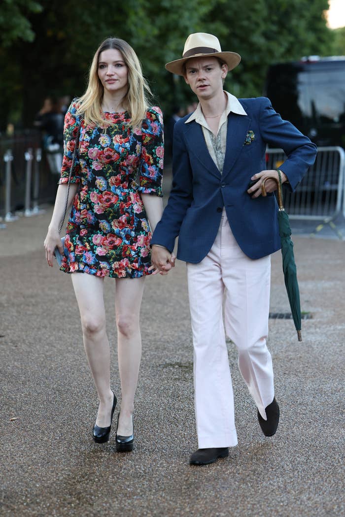 Thomas Brodie-Sangster and Talulah Riley walking hand-in-hand. Talulah is wearing a short floral summer dress and Thomas is wearing a jacket, slacks, and a fedora