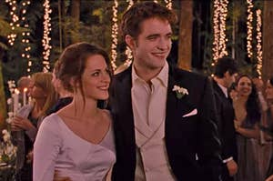 Edward and Bella from Twilight smiling together at their wedding reception, which is in a forest and features fairy lights on the trees