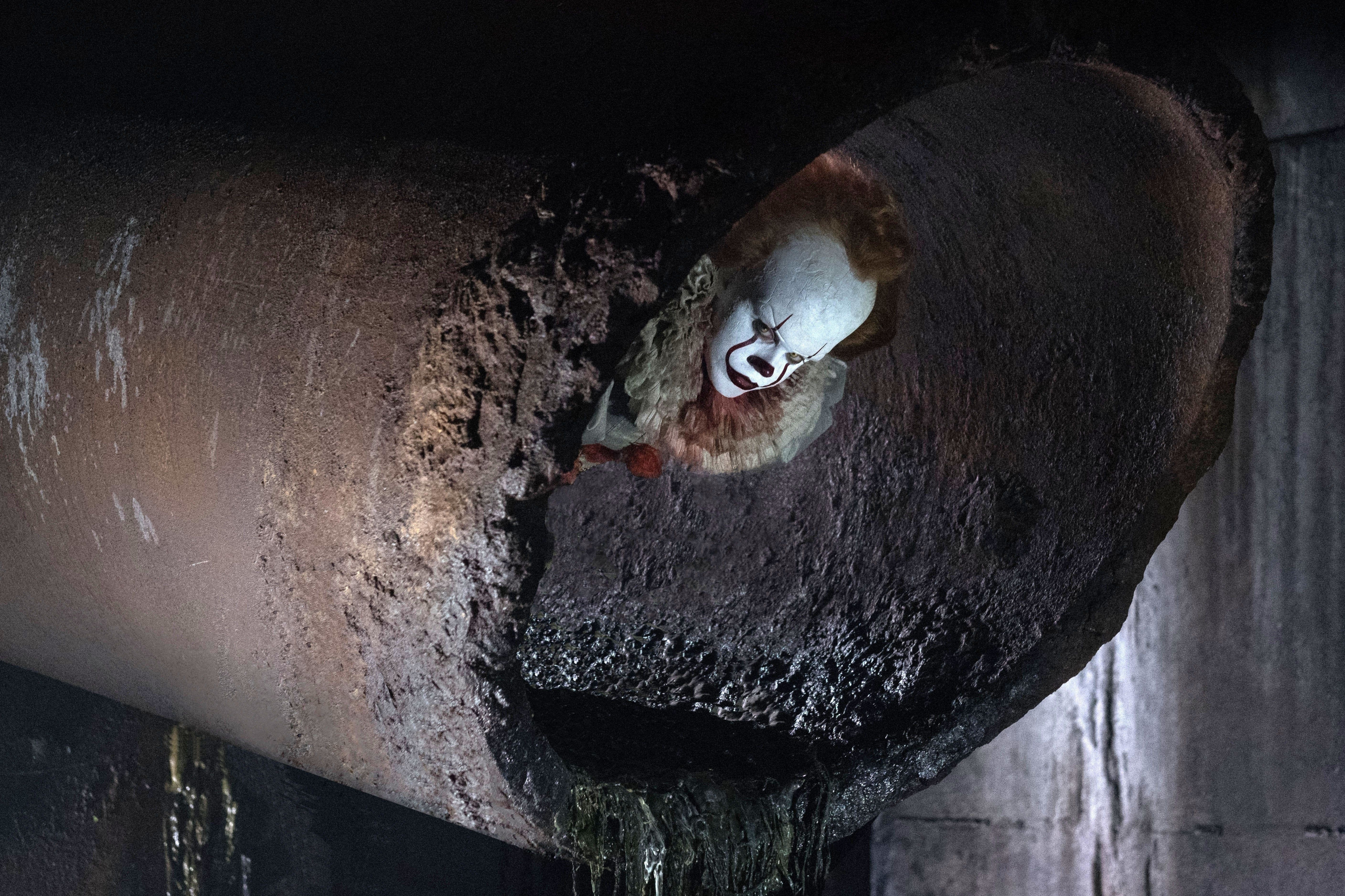 A terrifying clown sticks his head out of a rusty drain pipe