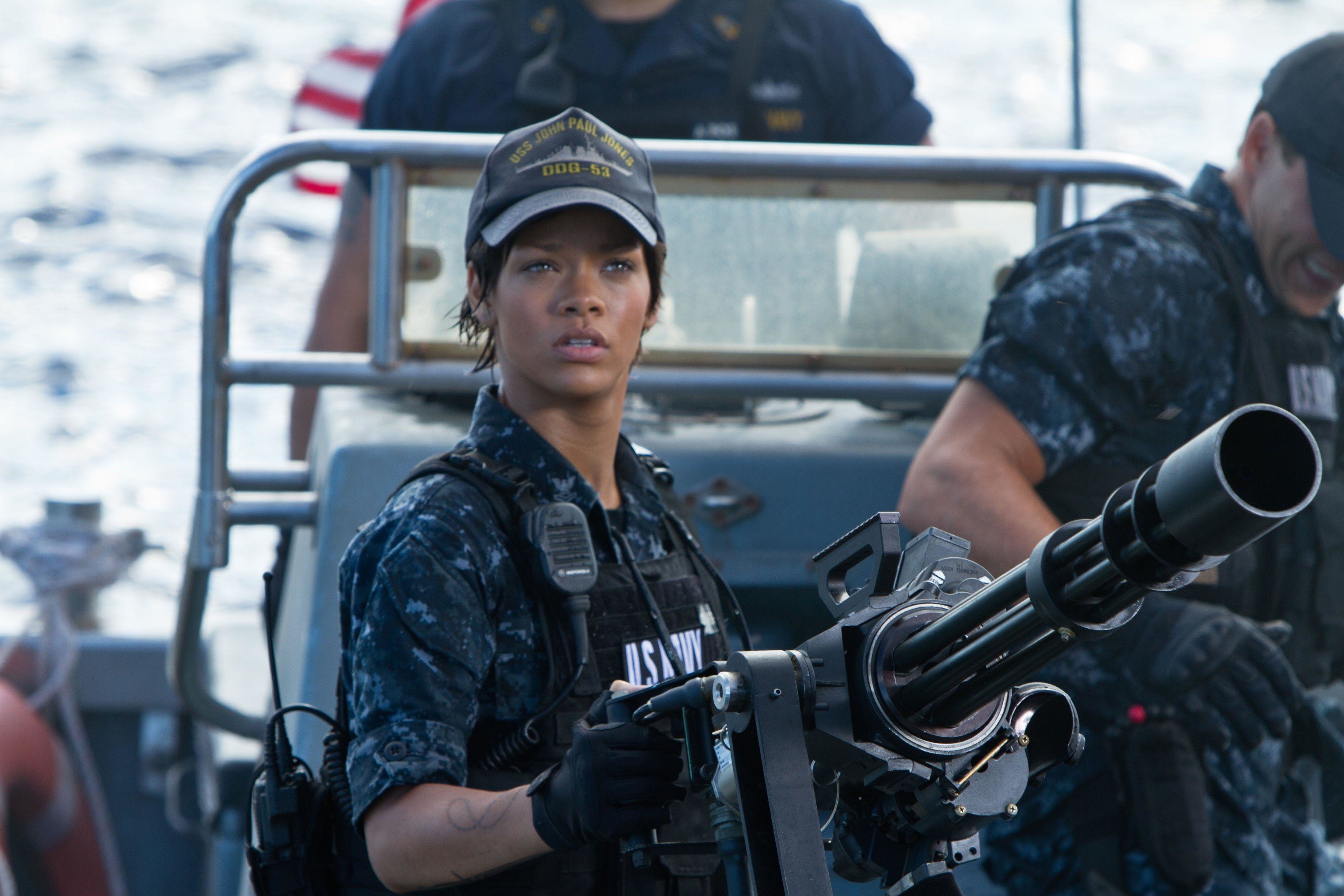 Rihanna stands behind a mini-gun and stares with concern on a speedboat