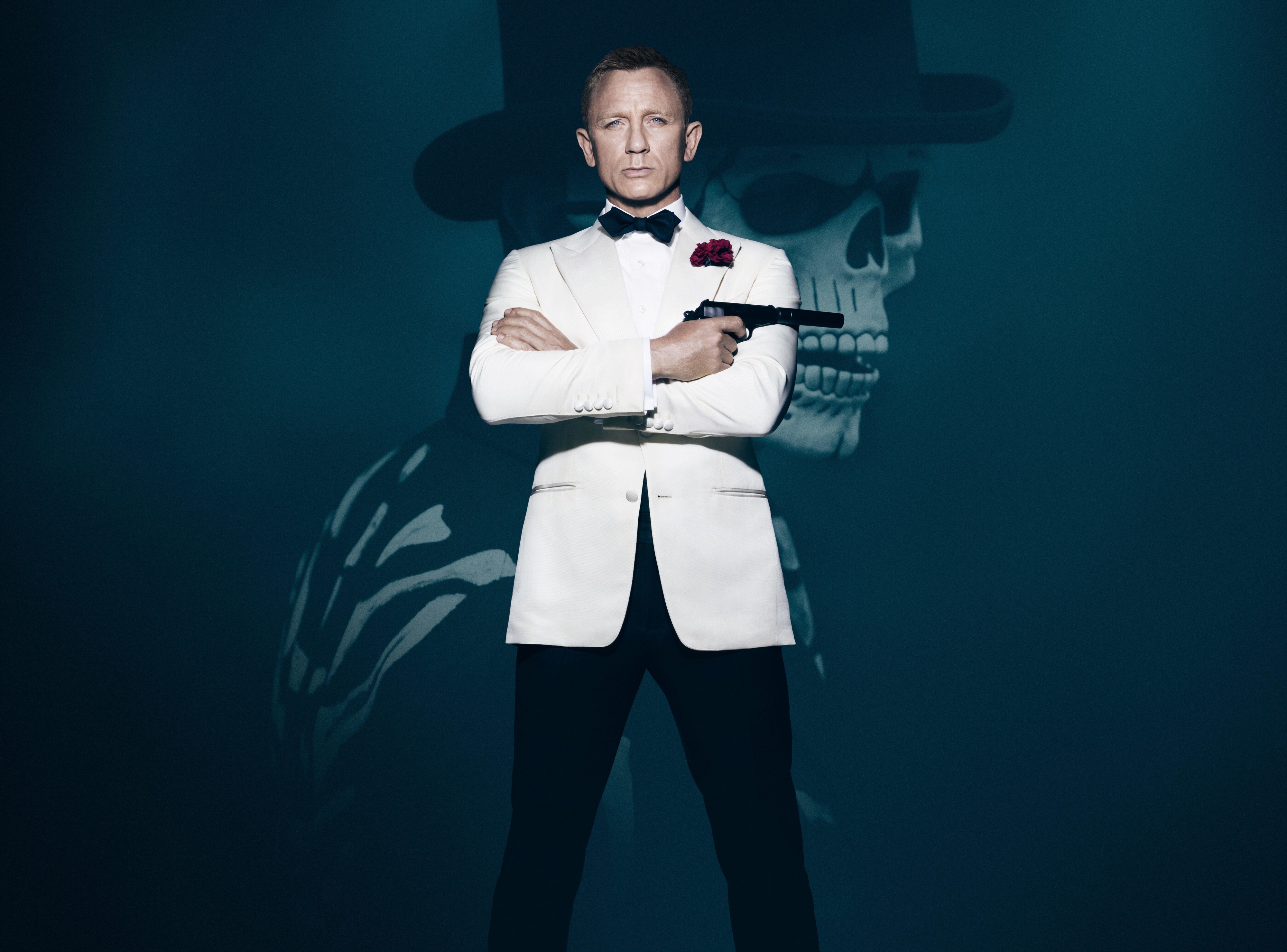 Daniel Craig poses in a white tuxedo with a pistol in front of the image of a man in a skull mask