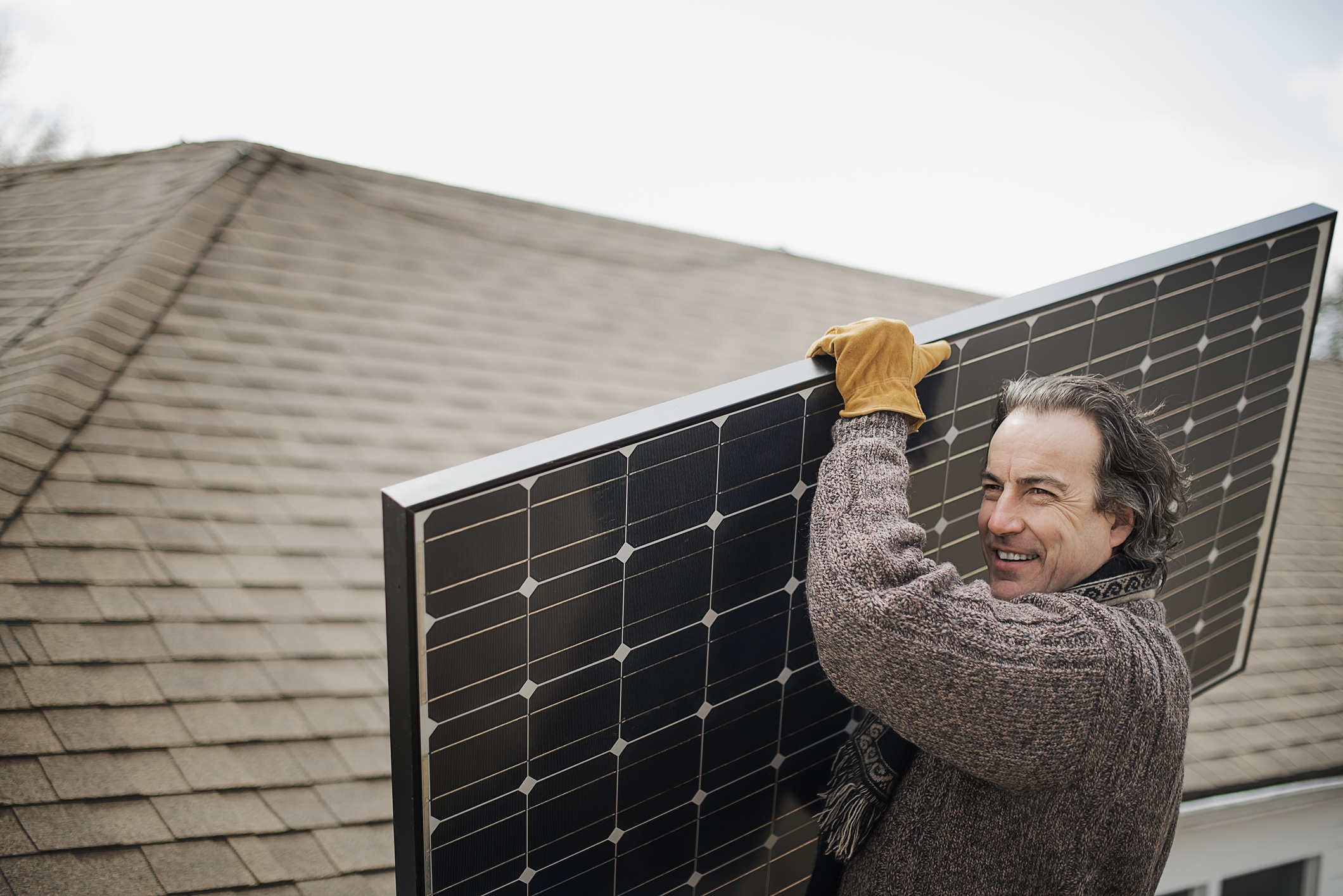A man in a sweater carries a solar panel on a roof