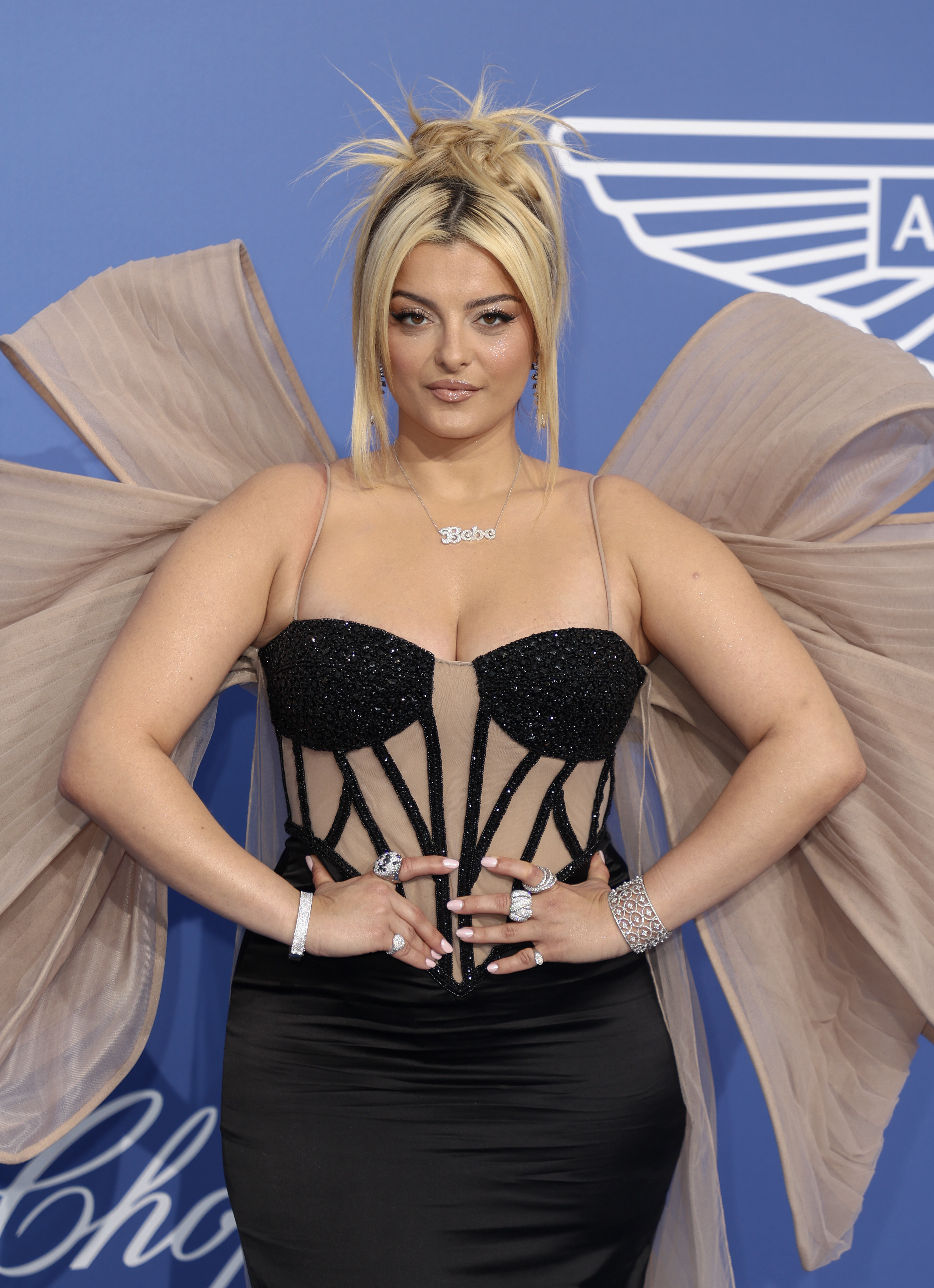 Bebe poses for photographers with her hands on her waist at a media event; she is wearing a corseted dress with a large bow on the back