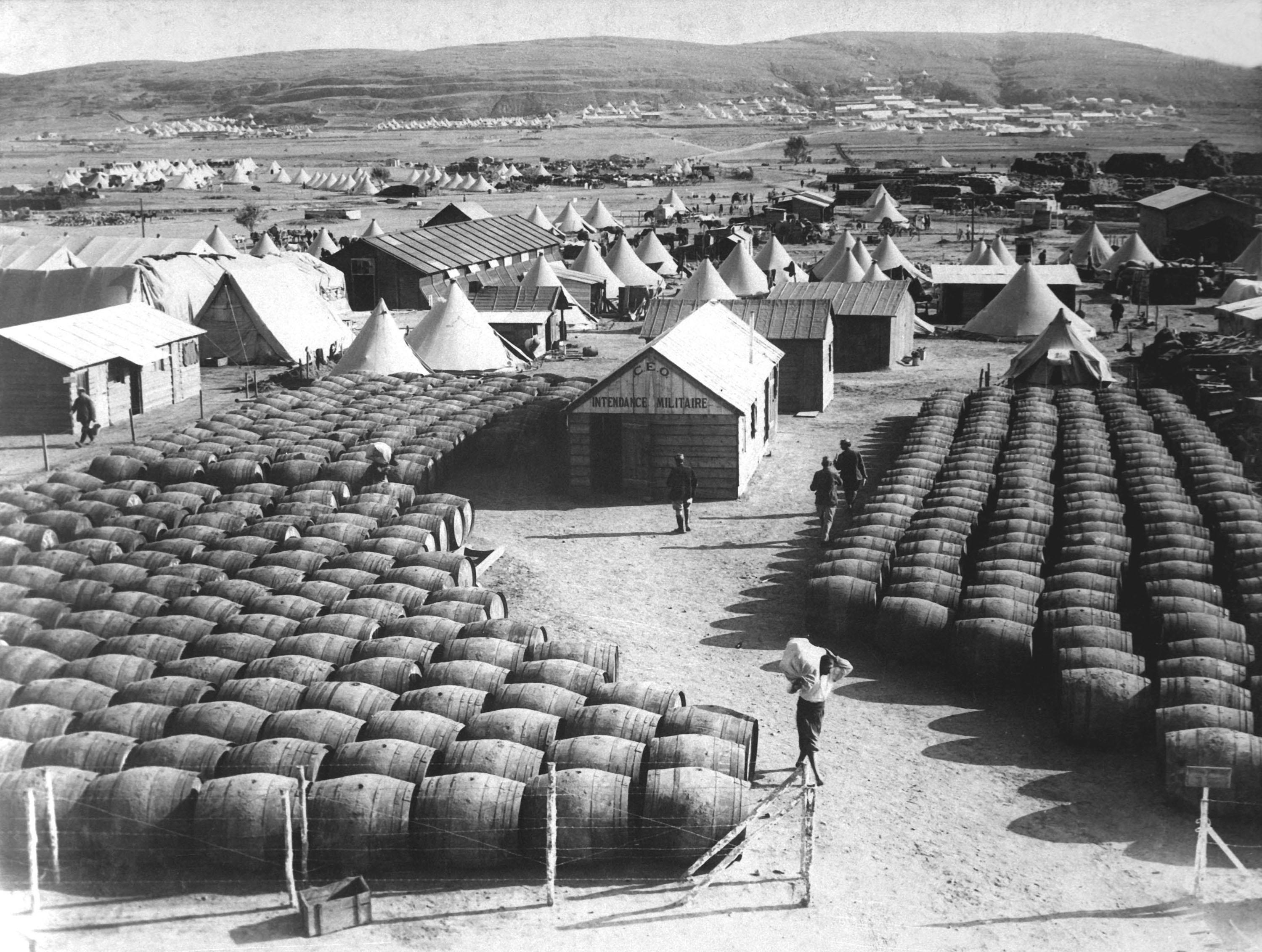 Hundreds of barrels on unpaved, grassless ground in a barren, open space with small, cone-shaped edifices nearby