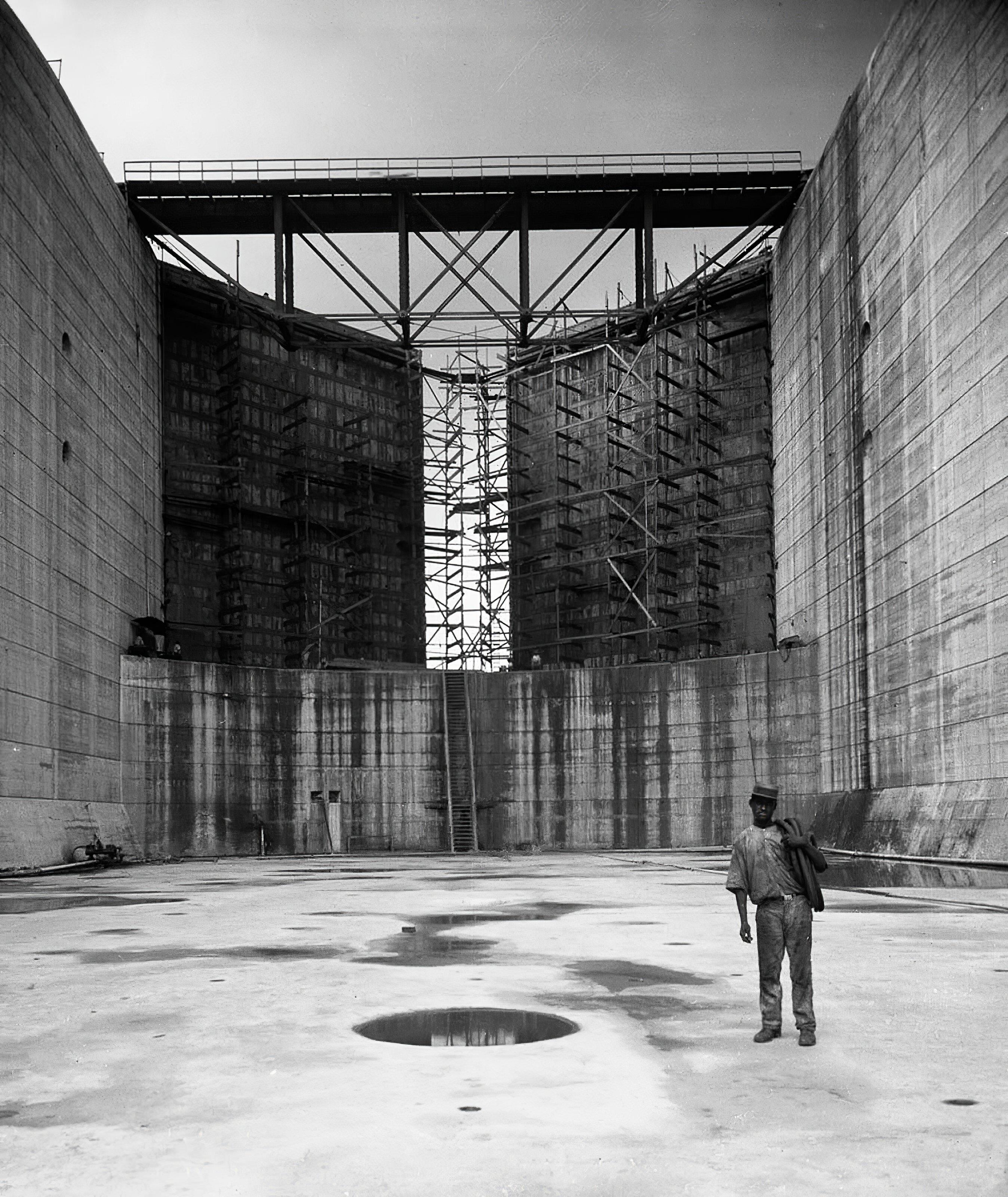 A solitary man standing in a cavernous empty space surrounded by very high walls with scaffolding and a bridge on top