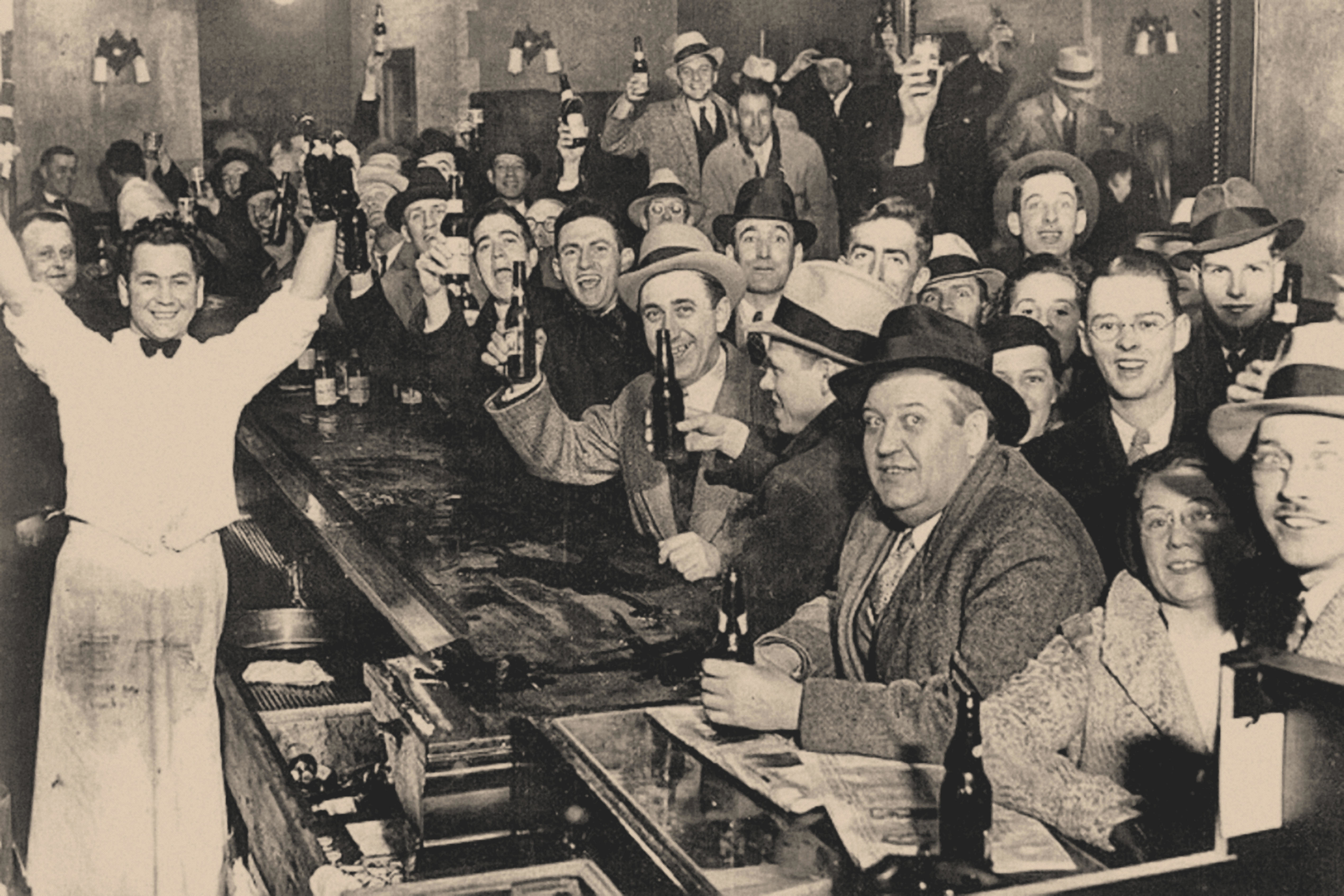 Smiling bartender with arms raised and a packed bar with smiling men, most holding beer bottles