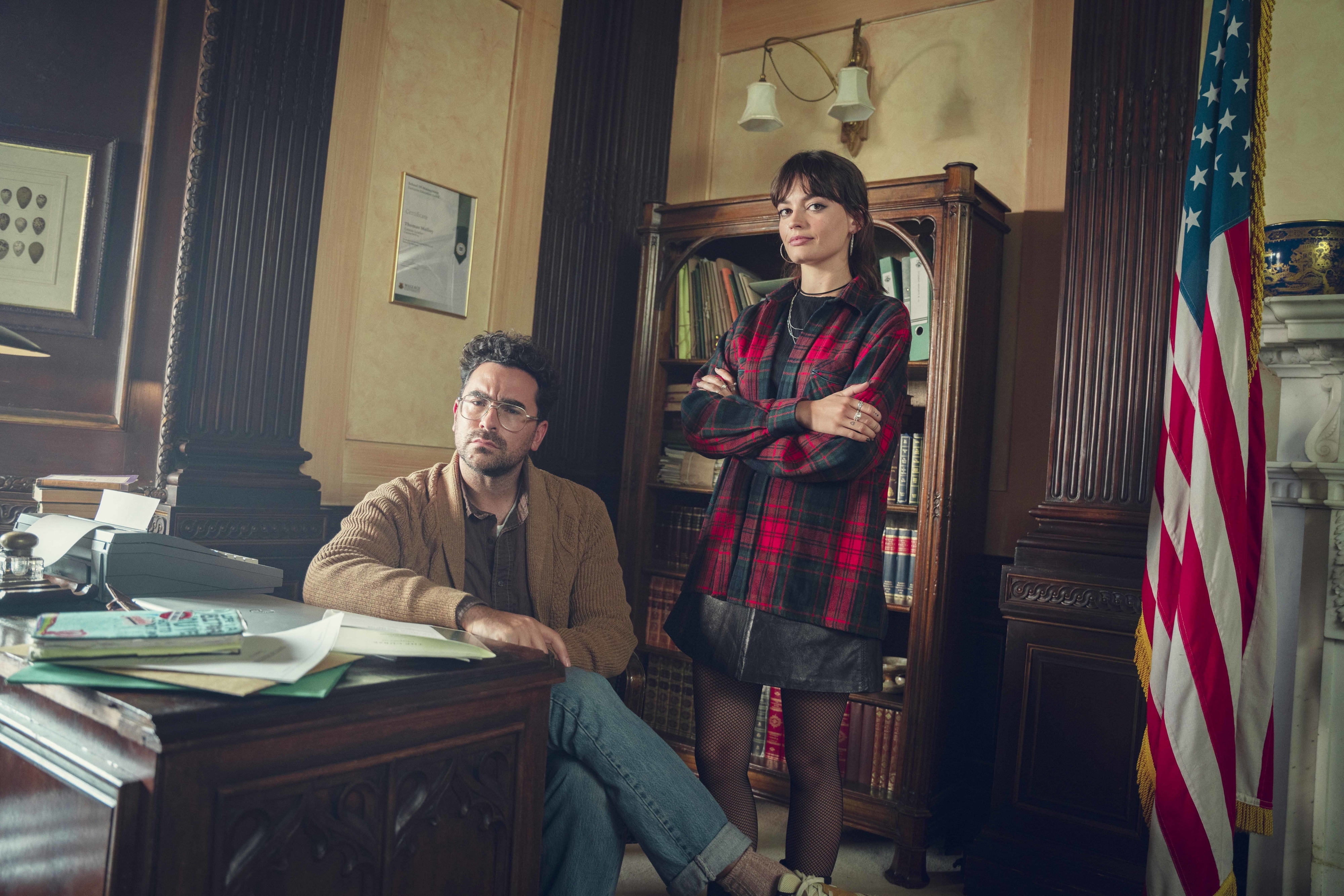 Dan sits at a desk, while Emma stands at his side with her arms folded