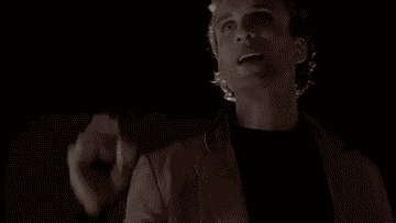 A person extends their hand while speaking animatedly at night, wearing a light jacket over a dark shirt