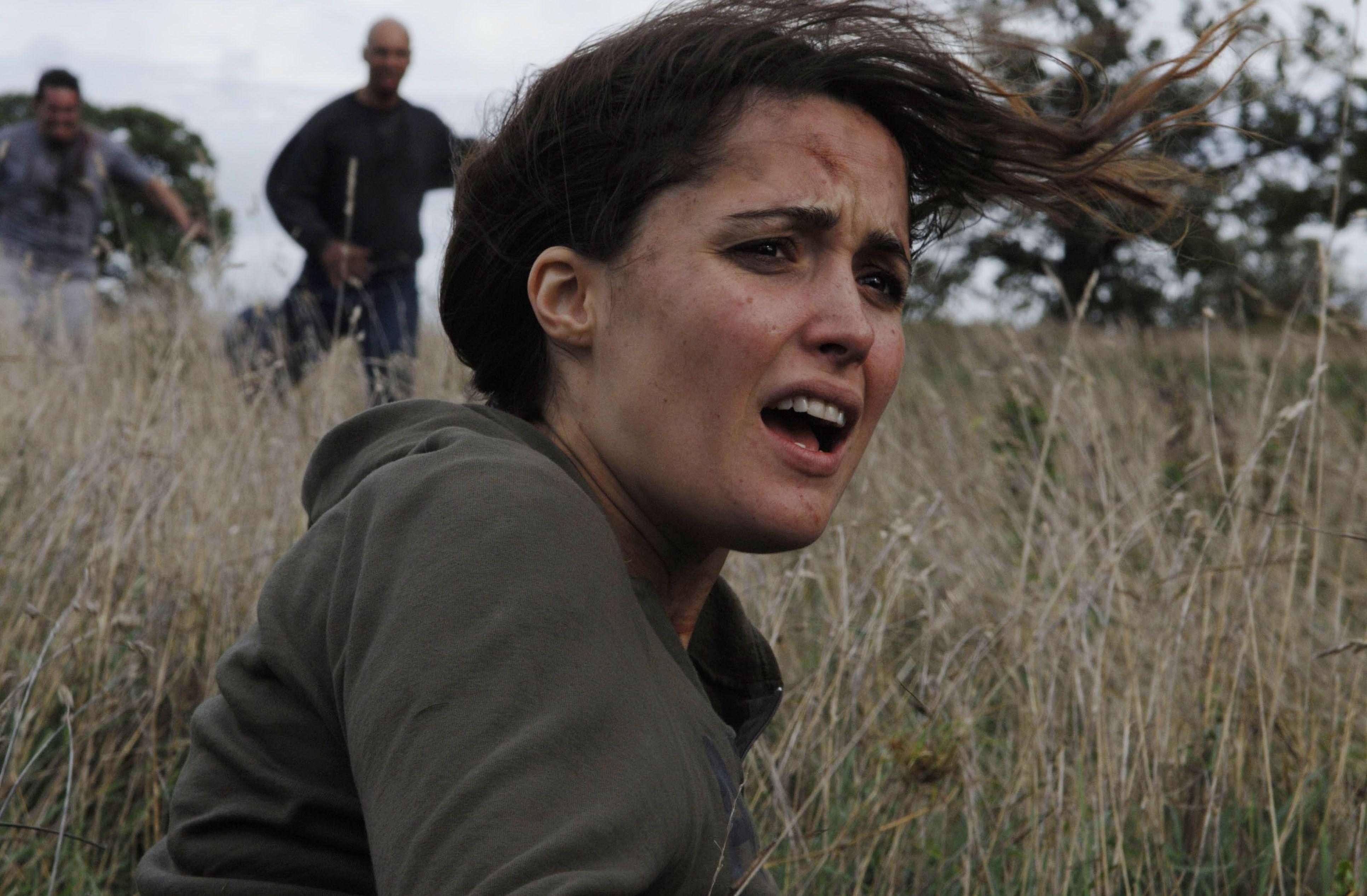 Rose Byrne sits in long grass while zombies run toward her