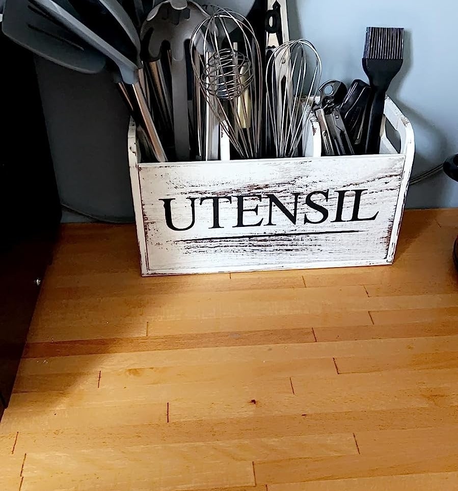 The white handled bin with UTENSIL in black font on front, filled with various cooking spoons, whisks, and other tools