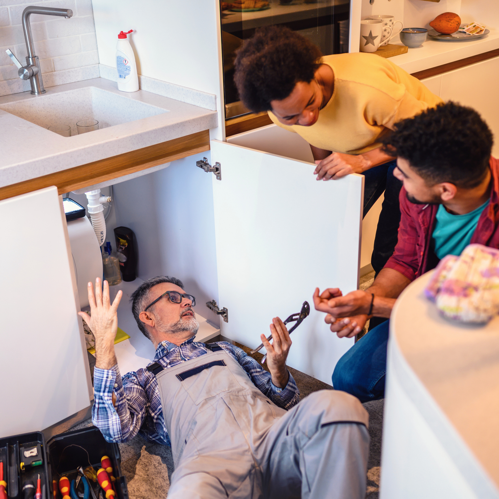 A plumber working and talking to a couple hovering over him