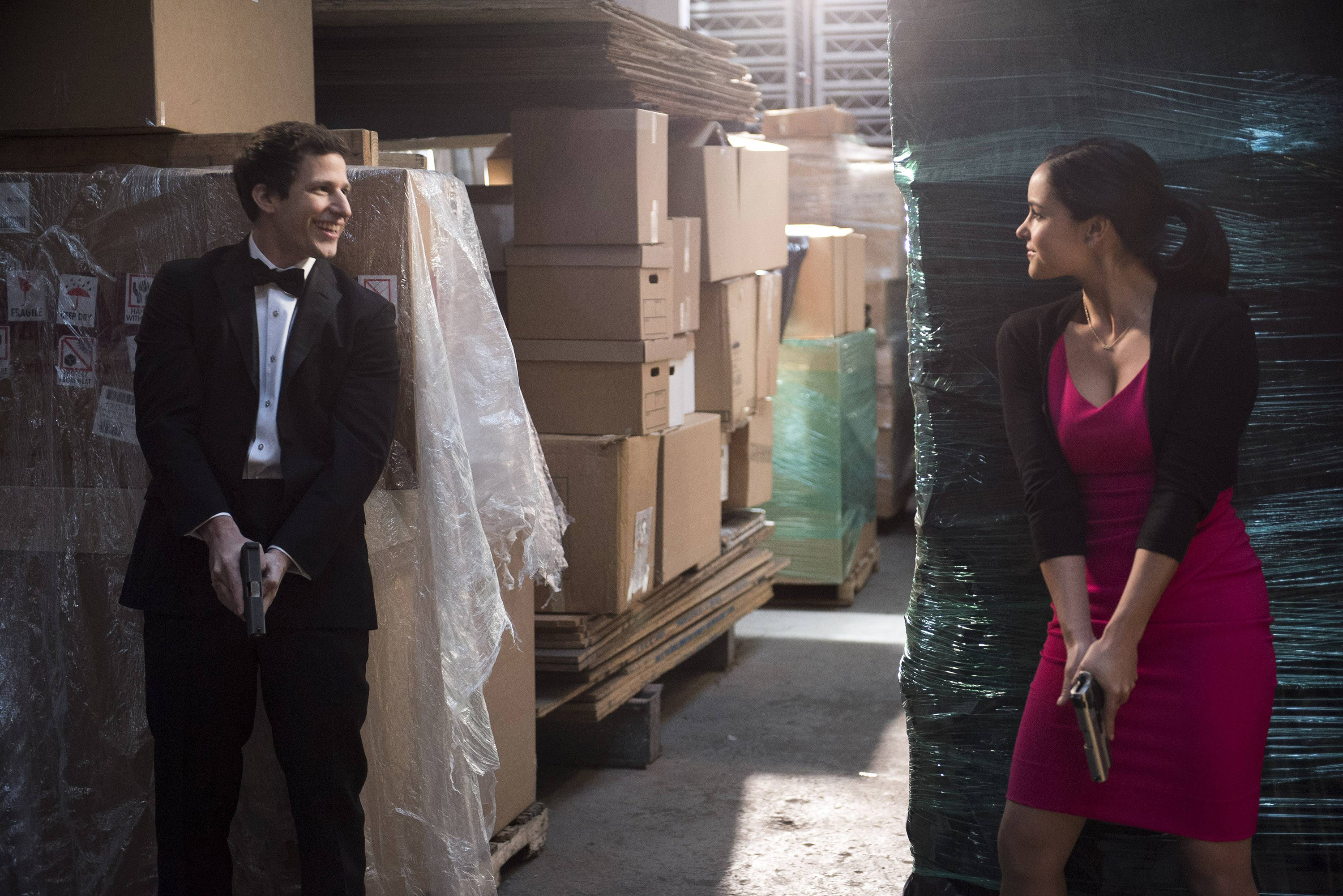 Andy Samberg and Melissa Fumero, dressed in fancy wedding clothes, hide in a warehouse with their guns drawn